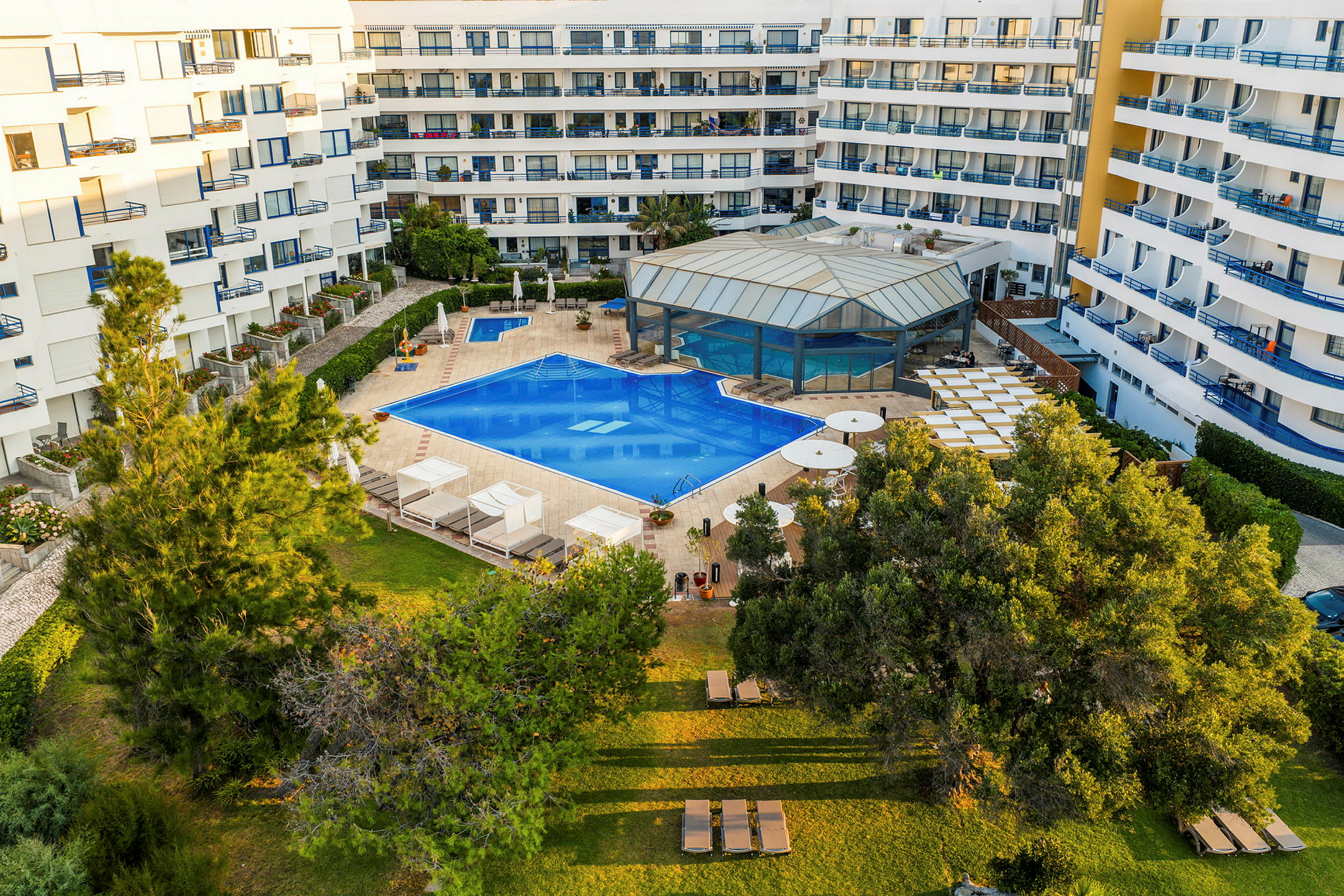 En bild som visar ett hotellområde med en stor utomhuspool omgiven av solstolar och grönska. Bakom poolen syns hotellets byggnad med flera balkonger. (AI-genererad text)
