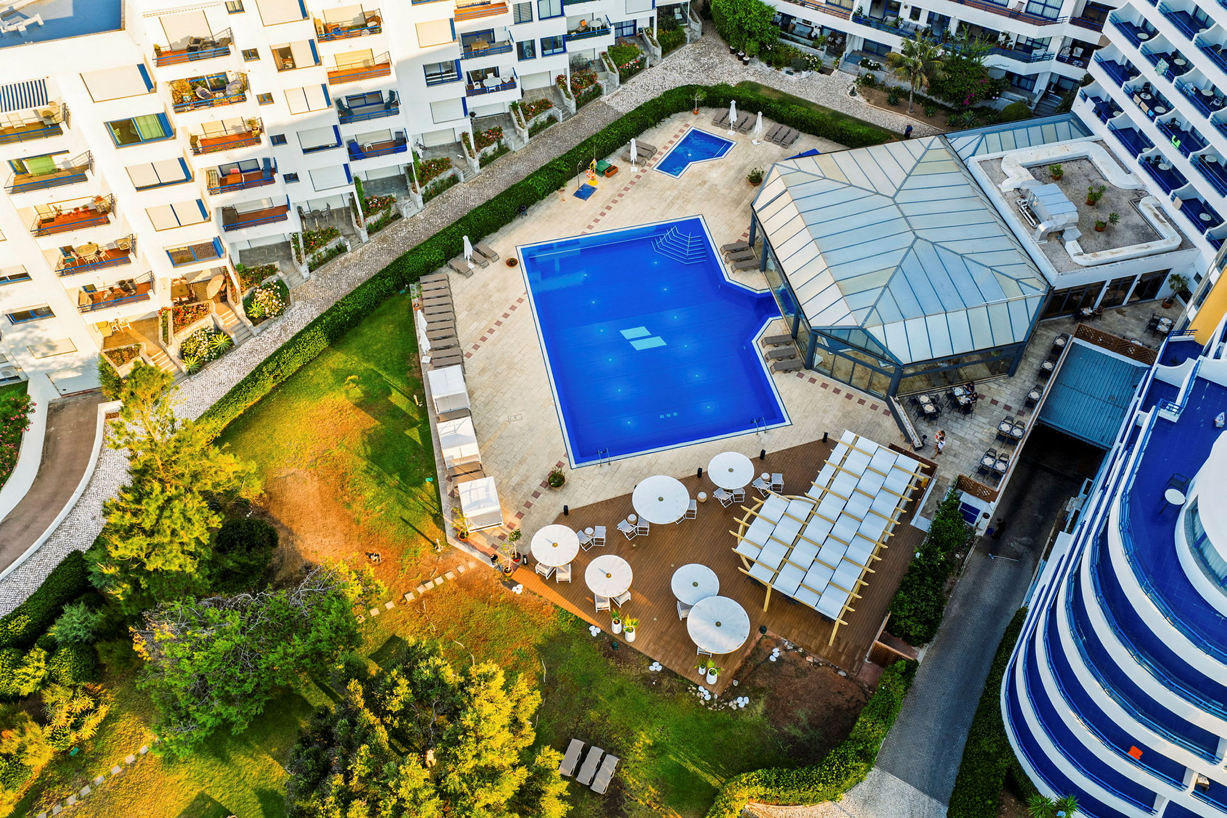 Flygfoto av ett hotellområde med en pool, solstolar, grönområden och uteservering. (AI-genererad text)