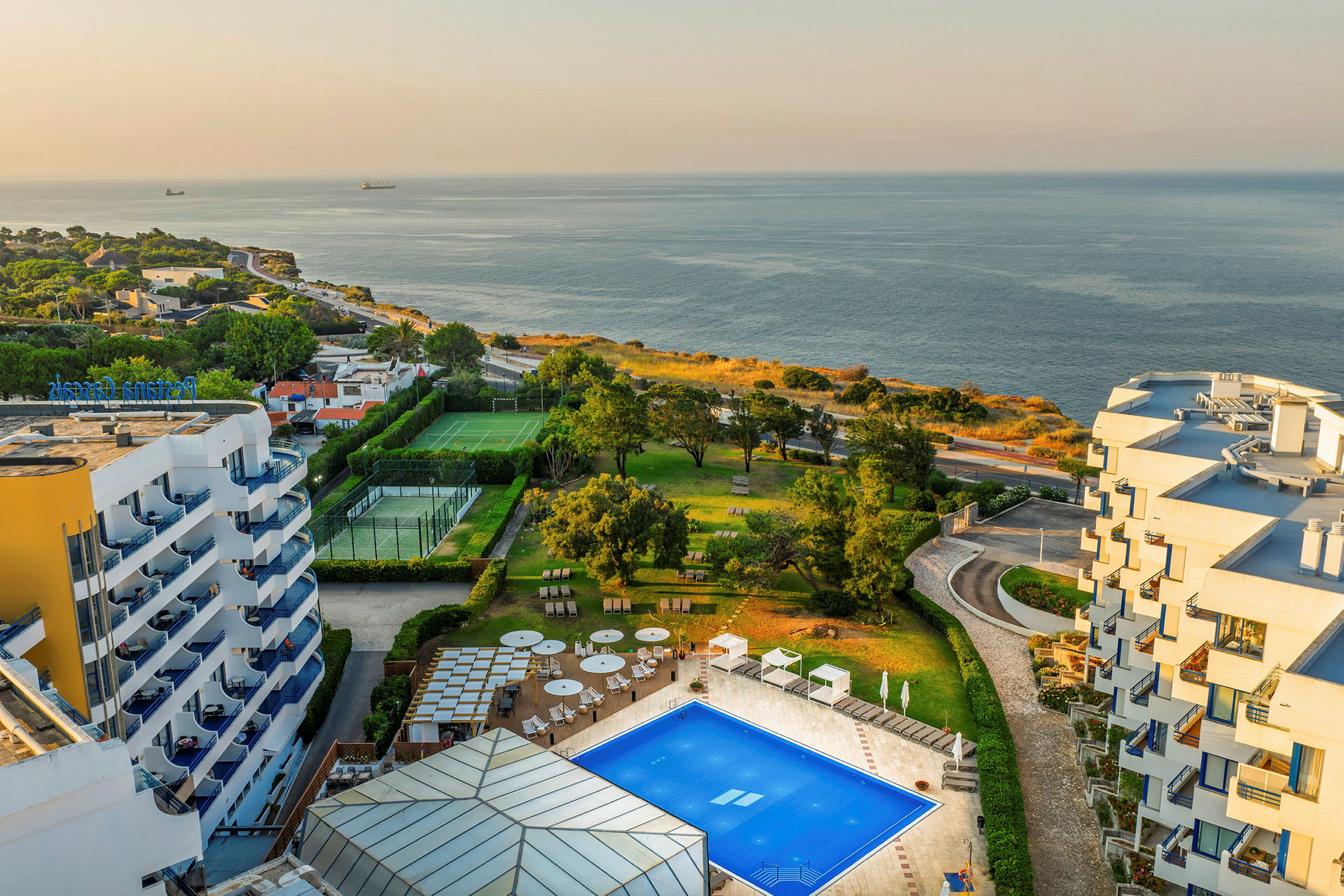 Utsikt över ett hotellområde vid havet med pool, grönområden, och sportplaner. (AI-genererad text)