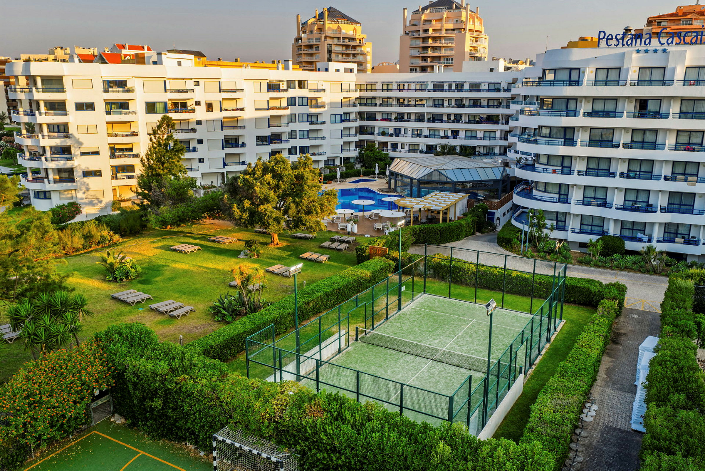En byggnad med vit fasad, ett poolområde med solstolar och pergola samt en tennisbana omgivet av grönområden och buskar. (AI-genererad text)