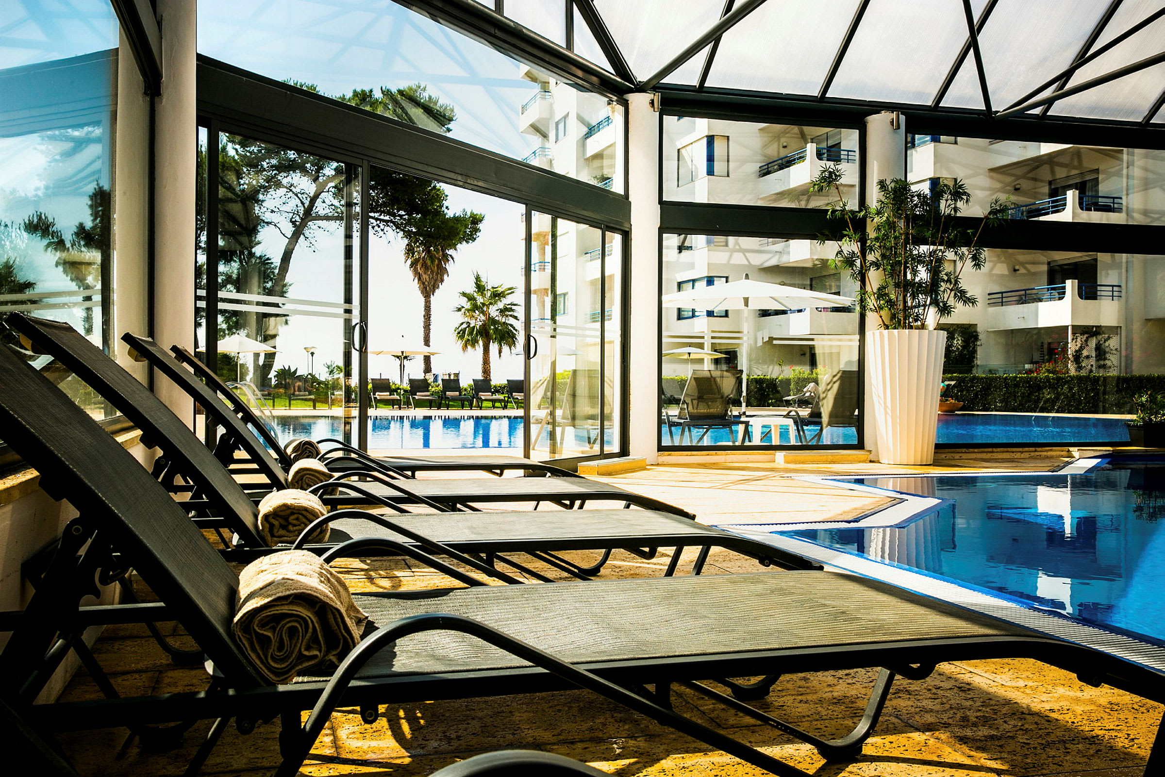 Solstolar i ett inomhusområde vid en pool med utsikt mot en trädgård och byggnader utanför. (AI-genererad text)
