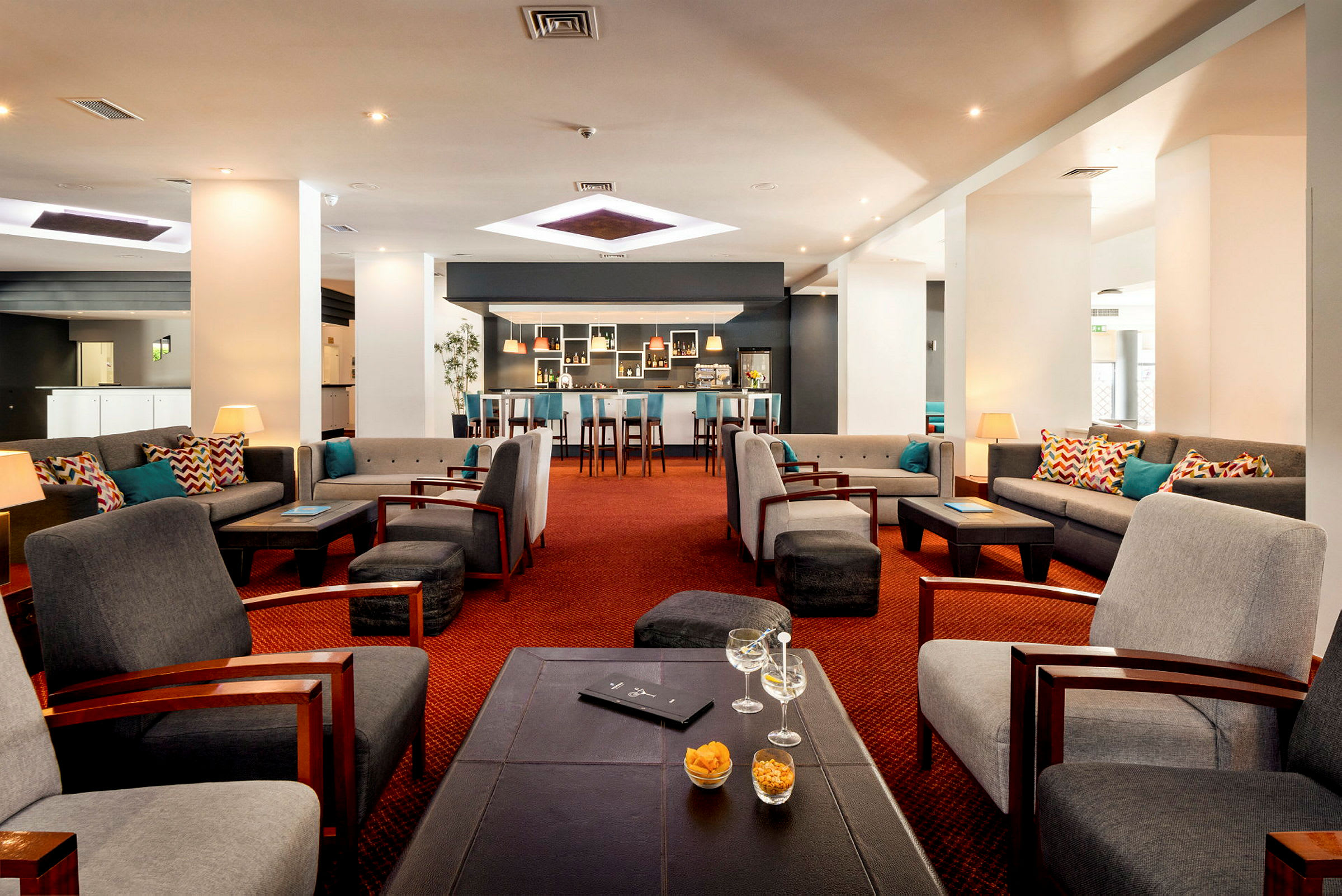 En lounge med bekväma sittmöbler, soffor och fåtöljer runt små bord. Väggar är vita och det finns en bardisk i bakgrunden. (AI-genererad text)