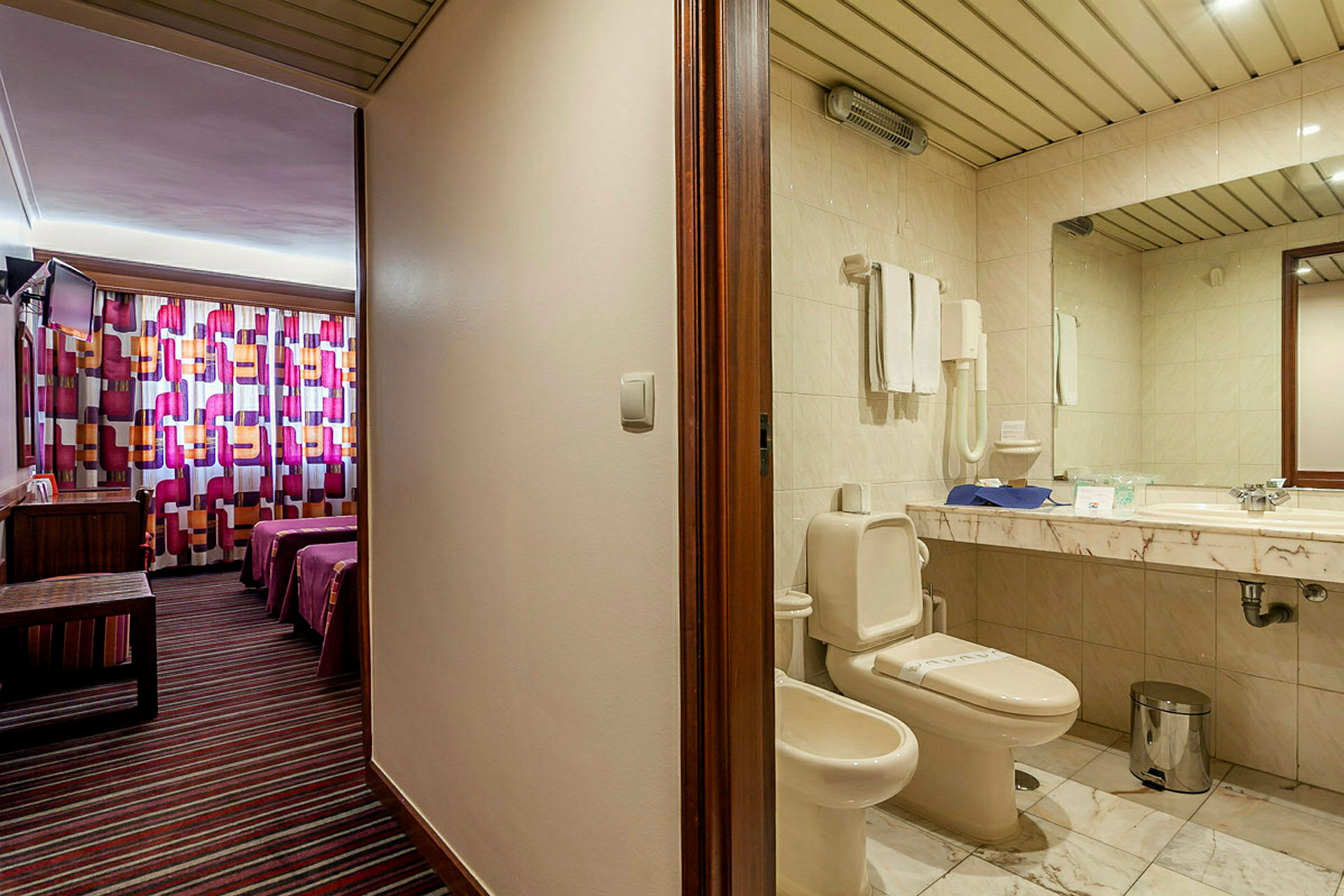 En hotellrumsinteriör som visar ett badrum med toalett och tvättställ till vänster, och ett sovrum med färgglada gardiner och två sängar till höger. (AI-genererad text)