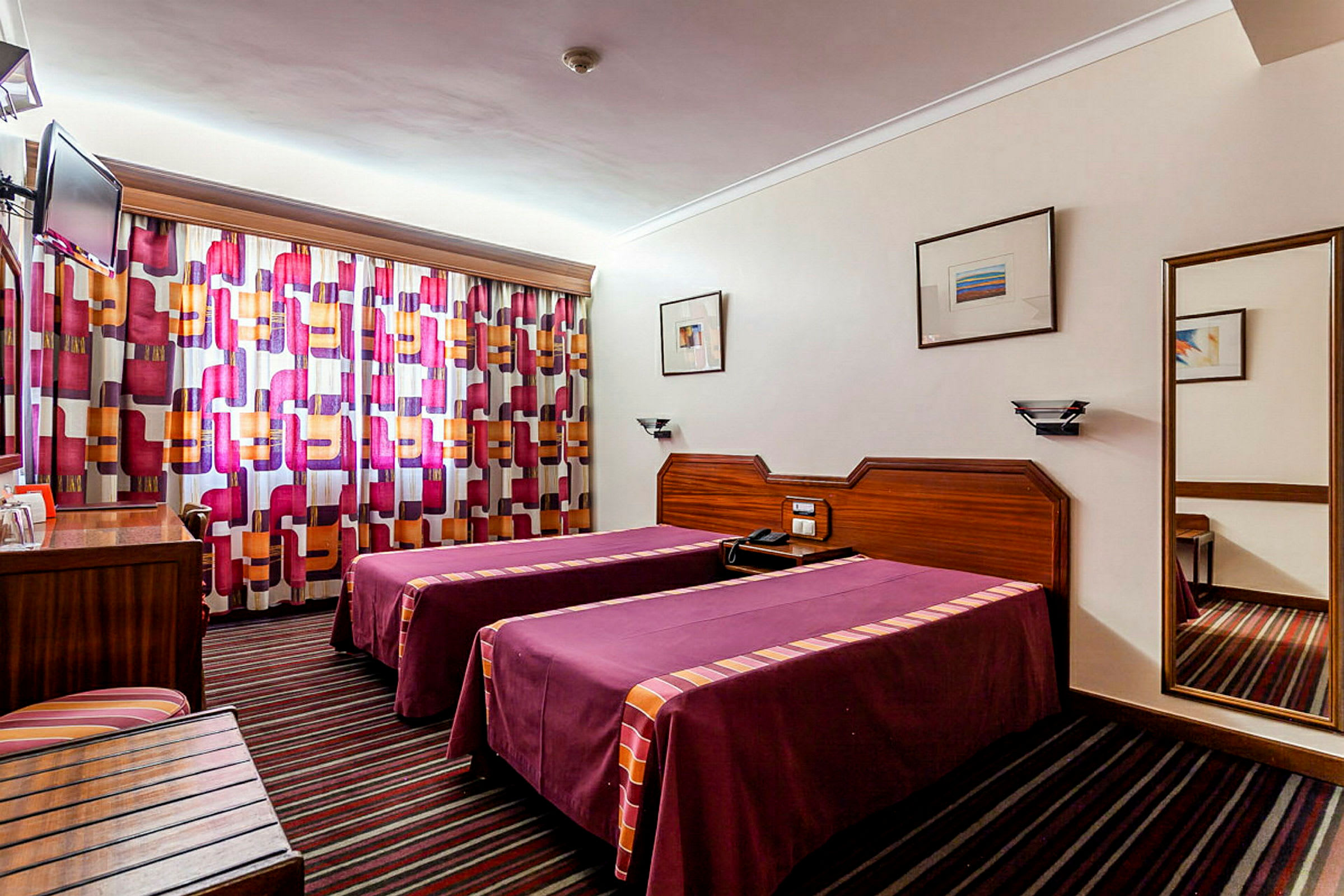 Hotellrum med två enkelsängar, färgglada gardiner och en väggmonterad spegel. (AI-genererad text)