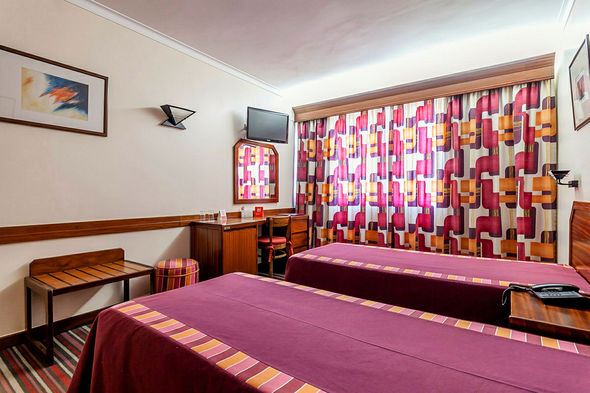 Ett hotellrum med två enkelsängar klädda i rödlila överkast. Rummet har färgglada gardiner och enkelt möblemang, inklusive skrivbord och stol. (AI-genererad text)