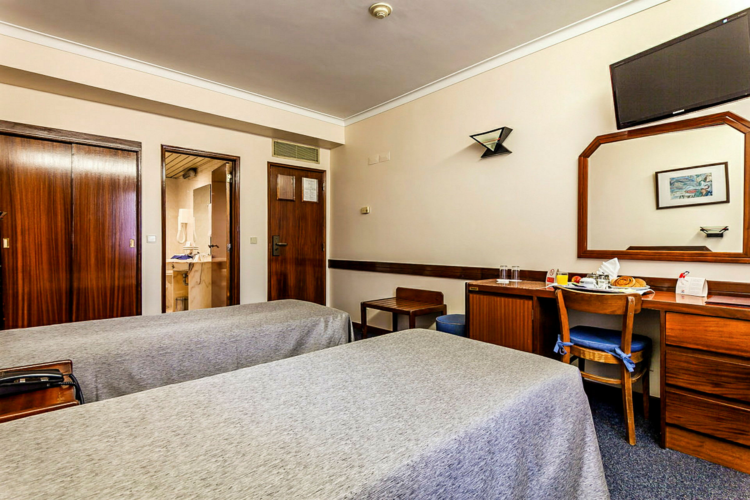 Ett hotellrum med två enkelsängar, en träskrivbord med en stol, en stor spegel och en väggmonterad TV. (AI-genererad text)