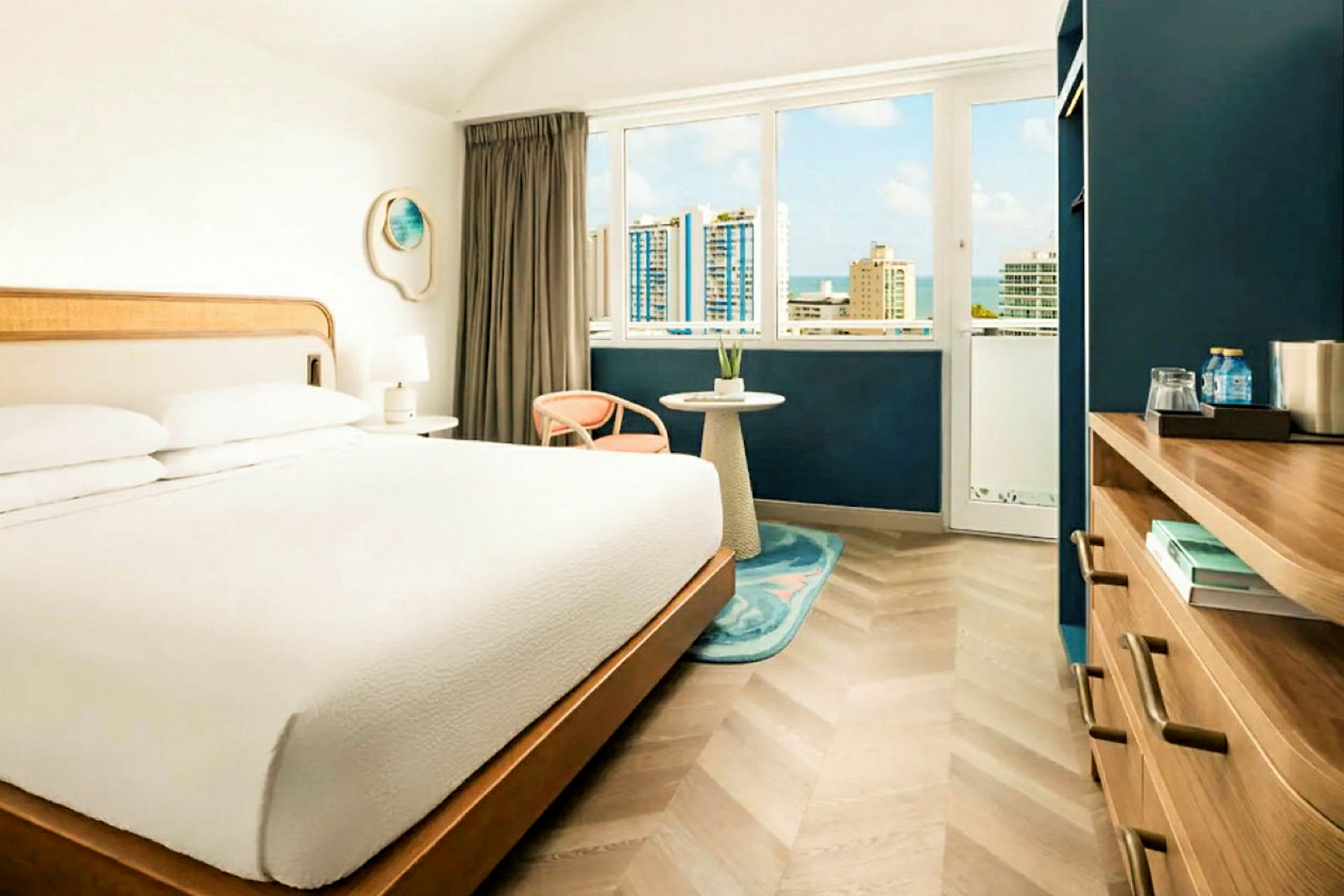 Ett hotellrum med en stor säng, ett fönster med utsikt över en stad med höghus och himmel, samt en möbel med vattenflaskor och böcker. (AI-genererad text)