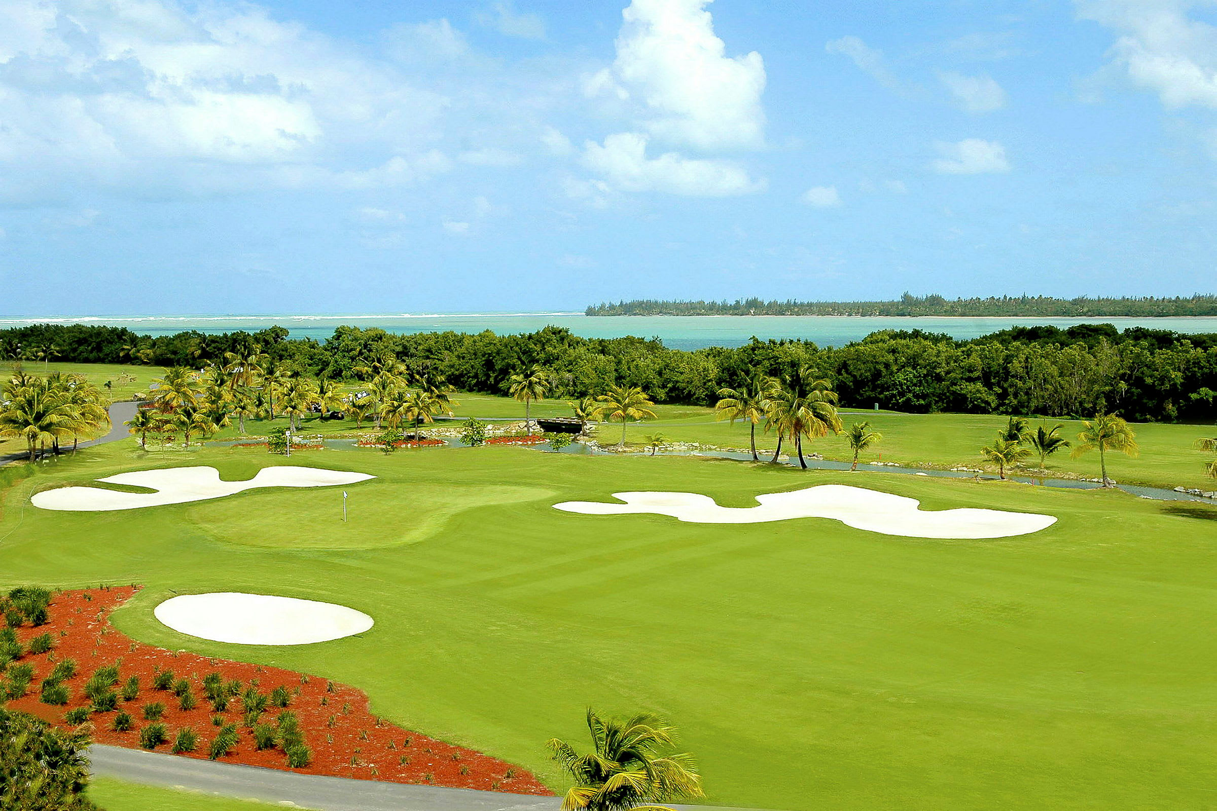 En golfbana med sandbunkrar och grön fairway, omgiven av palmer och med utsikt över havet. (AI-genererad text)