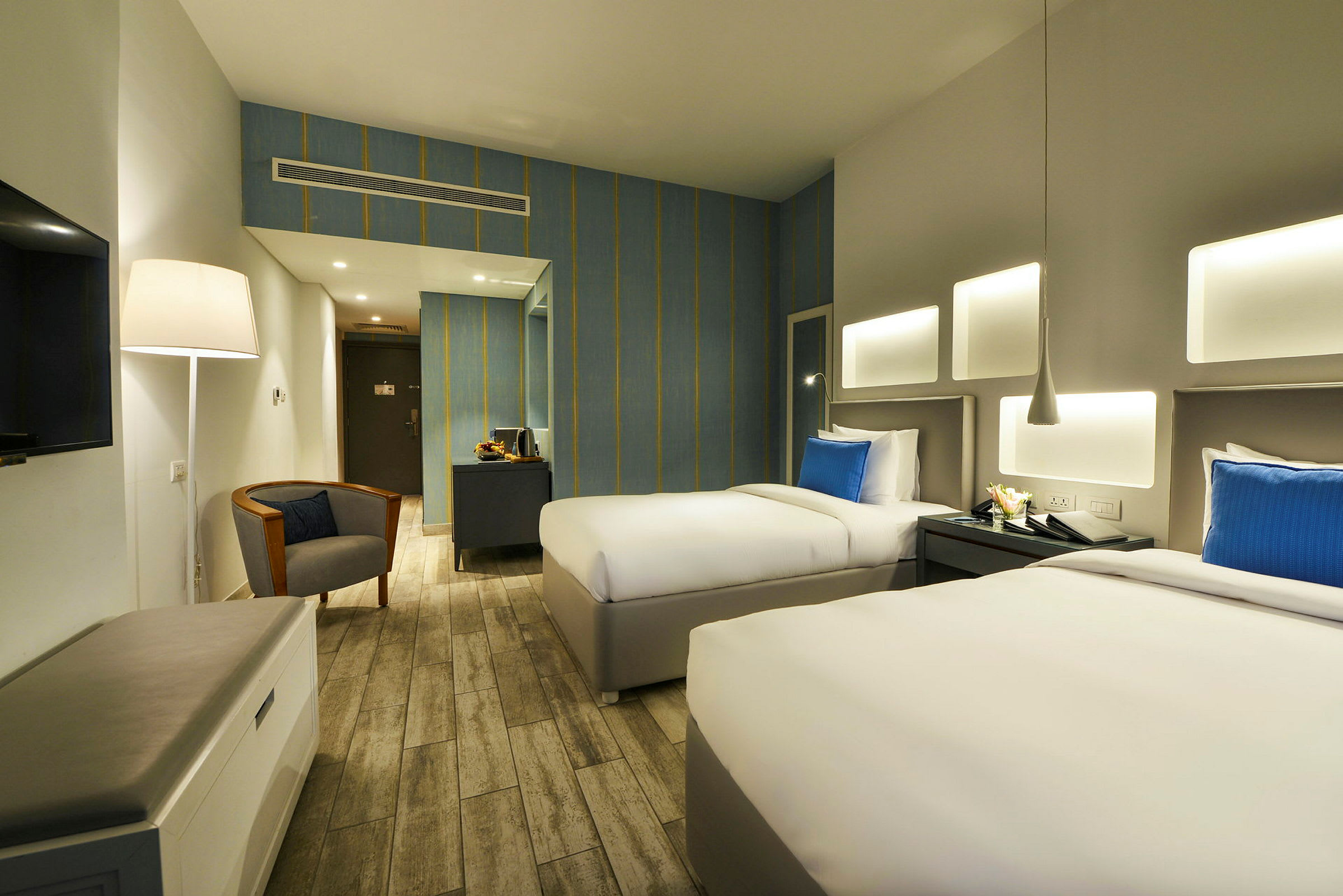 Ett hotellrum med två enkelsängar, blåa kuddar, ett skrivbord, en läsfåtölj och en lampa. (AI-genererad text)