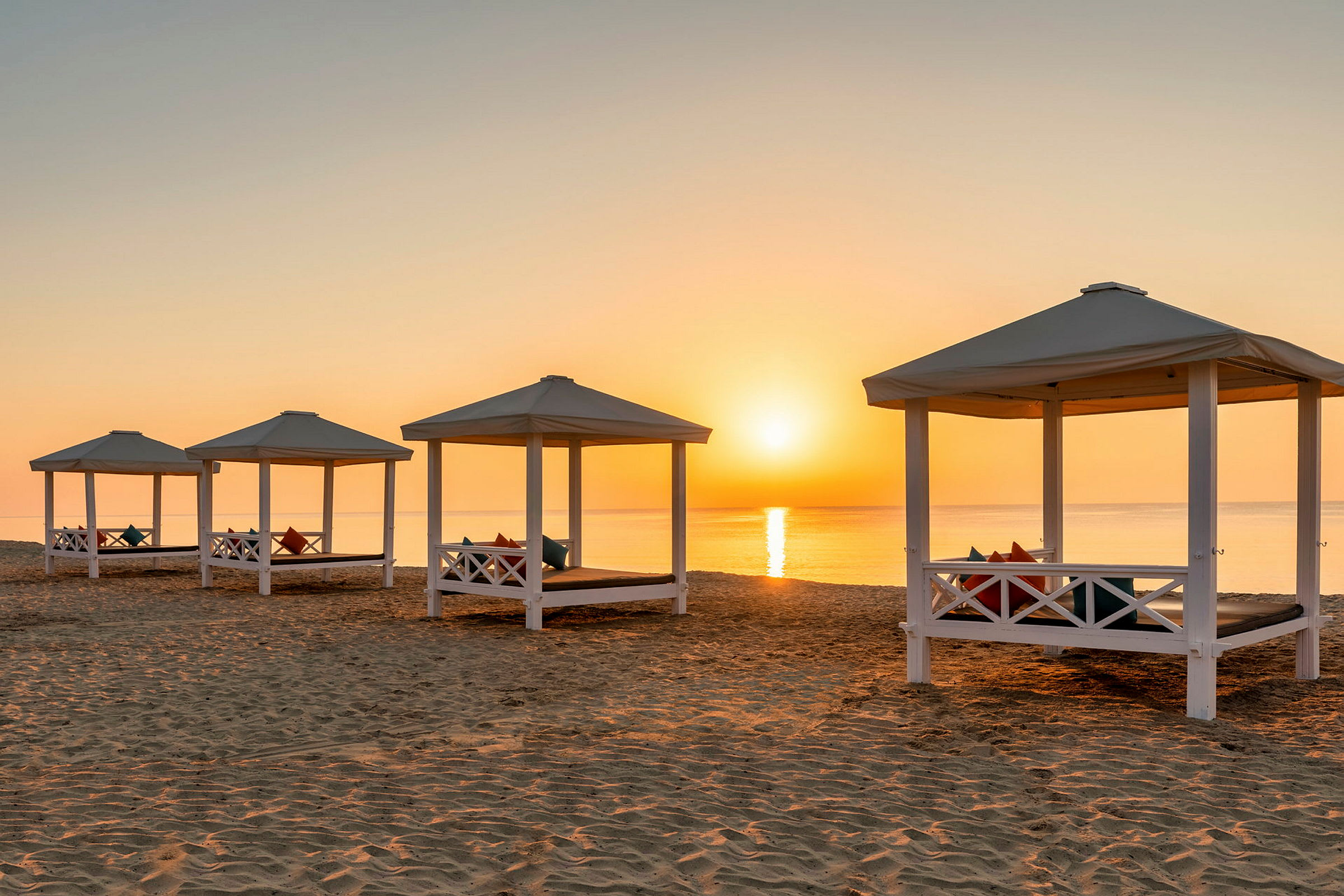 Bild av en strand med soluppgång. Flera vita byggnader med tak och kuddar ligger i sanden, och havet syns i bakgrunden. (AI-genererad text)