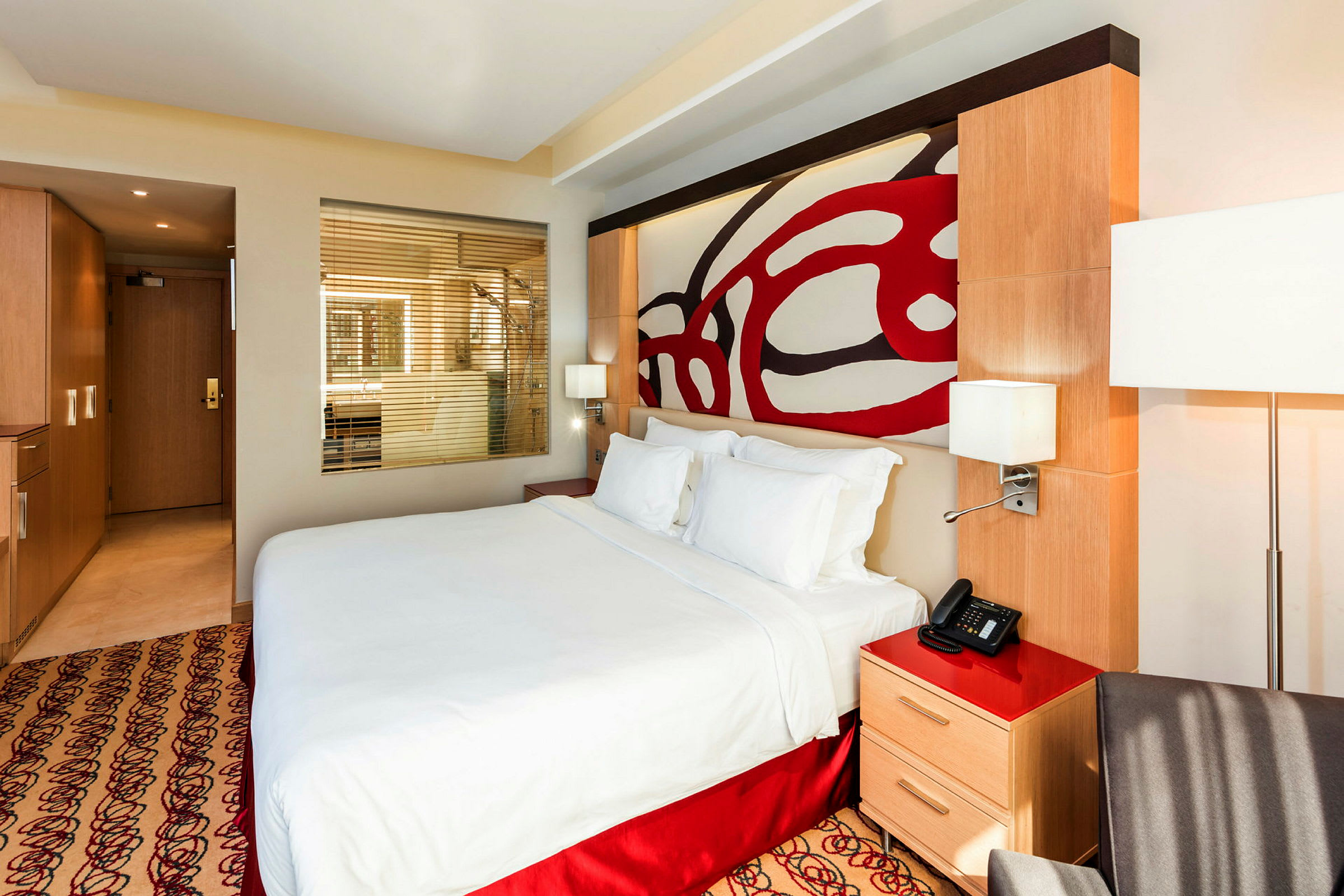 Ett modernt hotellrum med en stor säng och genomskinligt glasparti mot ett badrum. (AI-genererad text)