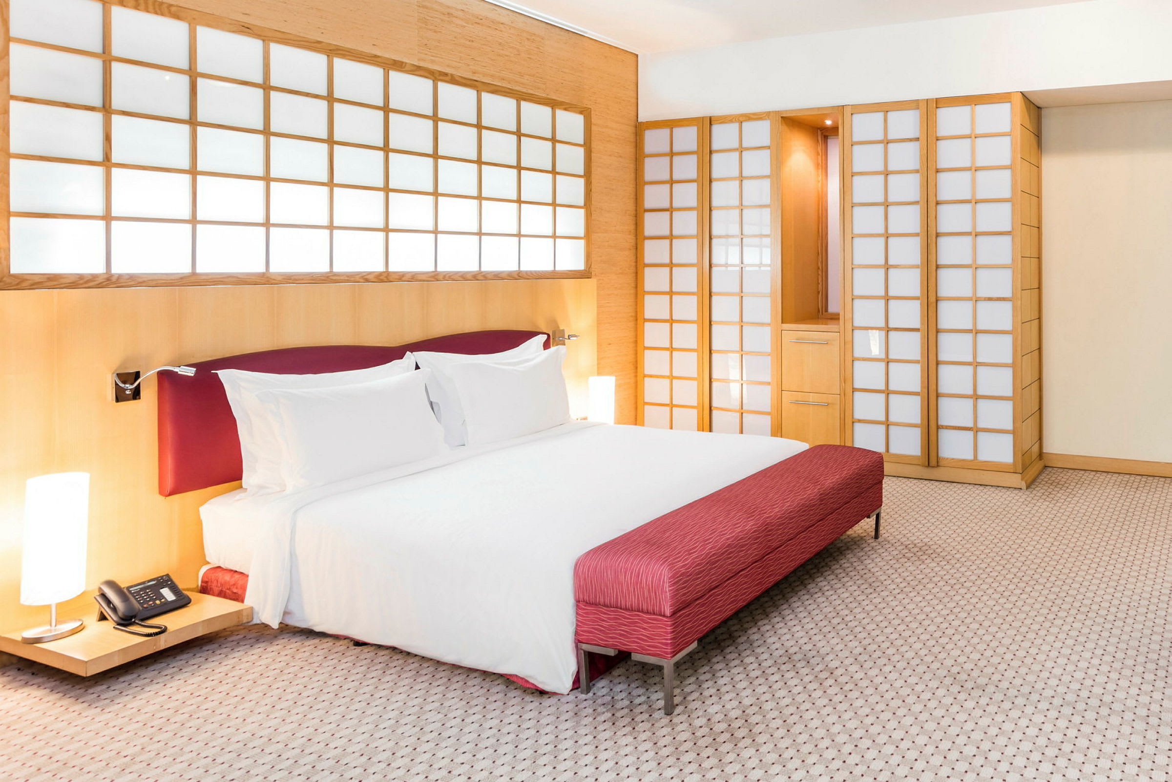 Ett hotellrum med en stor säng som har vita sängkläder och röda detaljer. Bakgrunden består av träpaneler och skjutdörrar med frostat glas. (AI-genererad text)