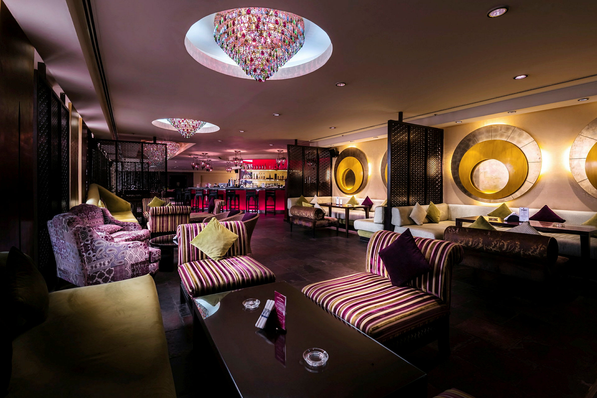 Ett elegant hotellbar med färgglada ljuskronor i taket, mjuka sittmöbler och soffor prydda med dekorativa kuddar. (AI-genererad text)