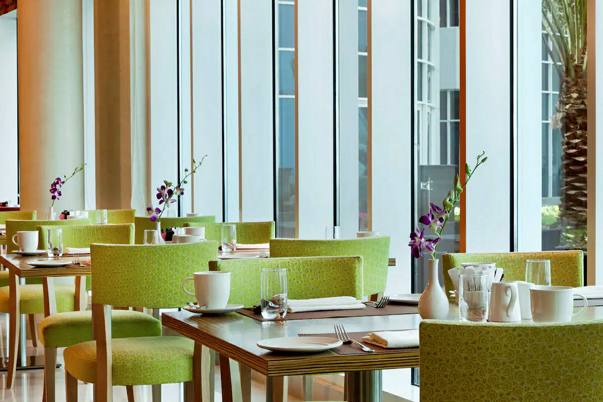 Ett modern restaurangområde med gröna stolar, uppdukade bord, vita koppar och lila orkidévaser. (AI-genererad text)