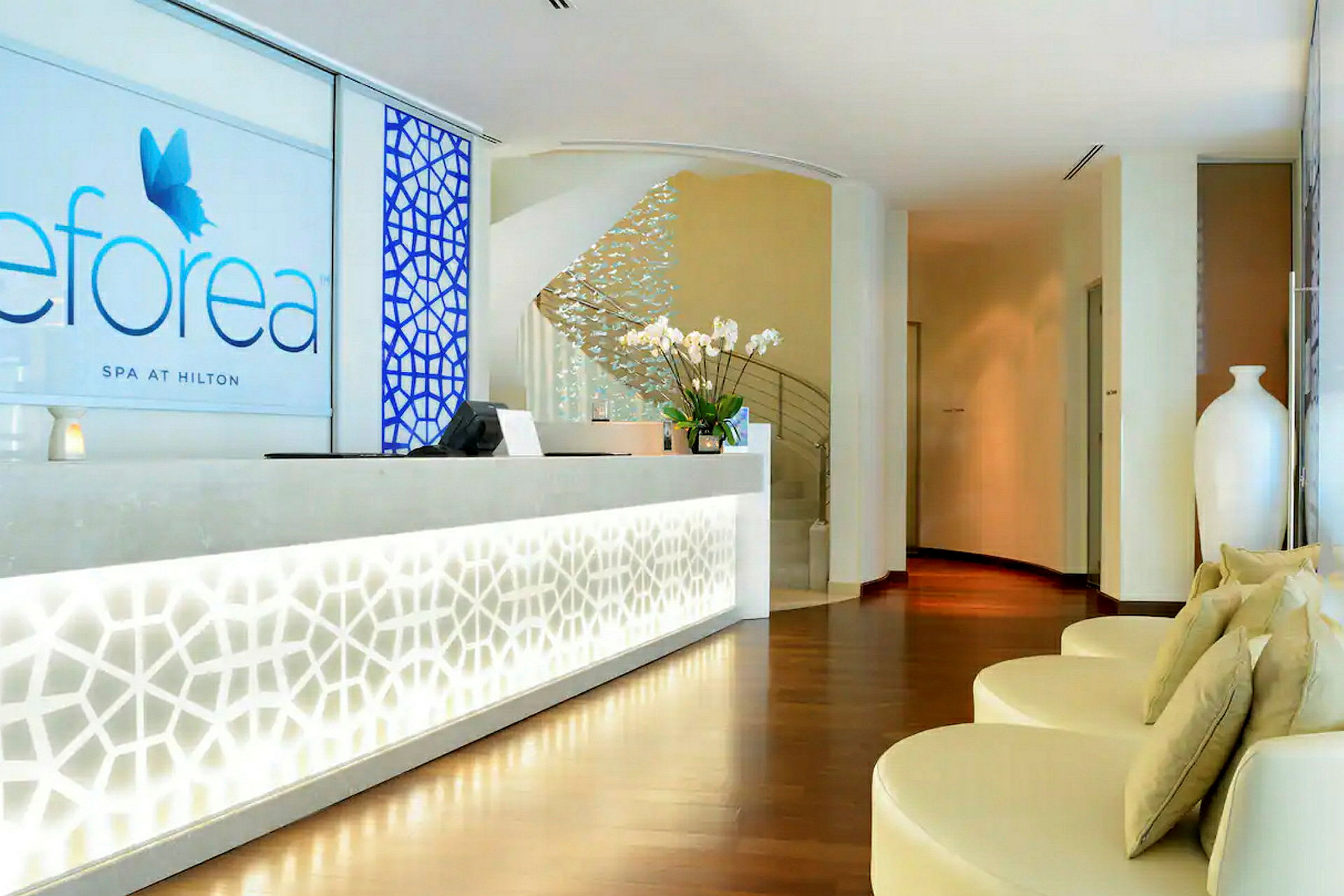 Bild av en receptionsdisk med modern vit design och spa-logotyp på väggen. I bakgrunden syns en trappa och dekorerade detaljer. (AI-genererad text)