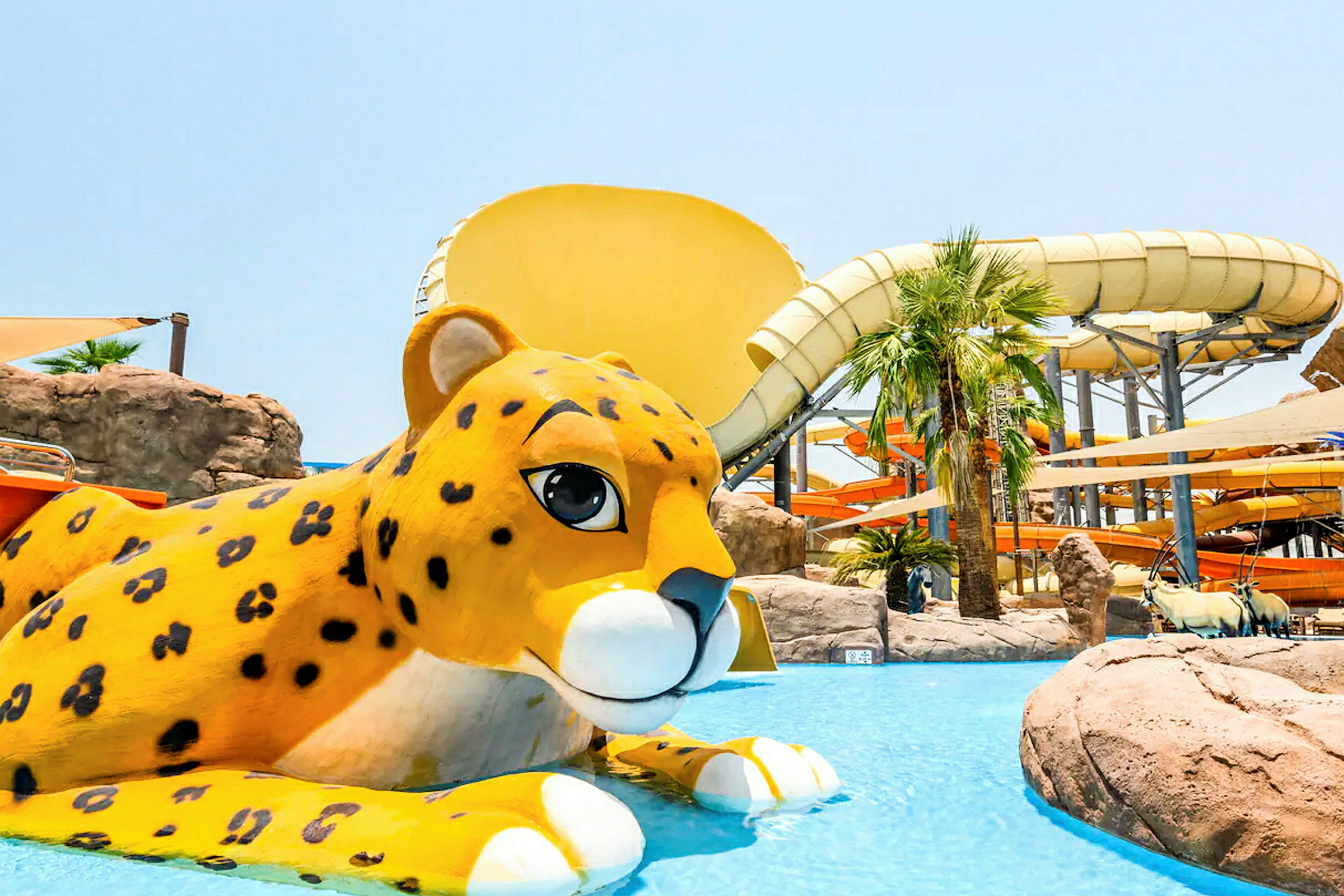 En gul leopardfigur i en pool framför en vattenpark med vattenrutschkanor. (AI-genererad text)