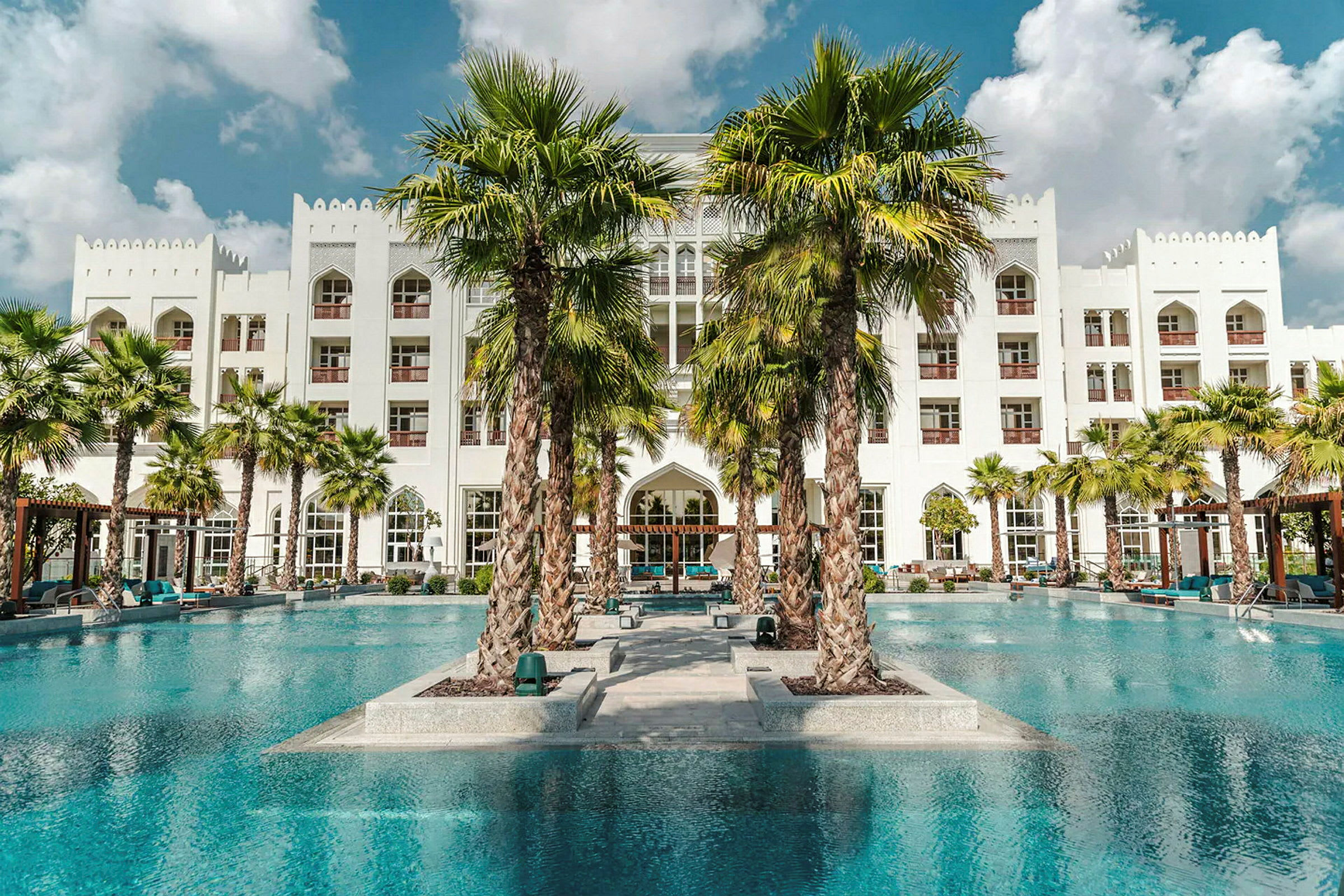 Ett modernt hotell med vit fasad sett från en pool, omgiven av palmer. (AI-genererad text)