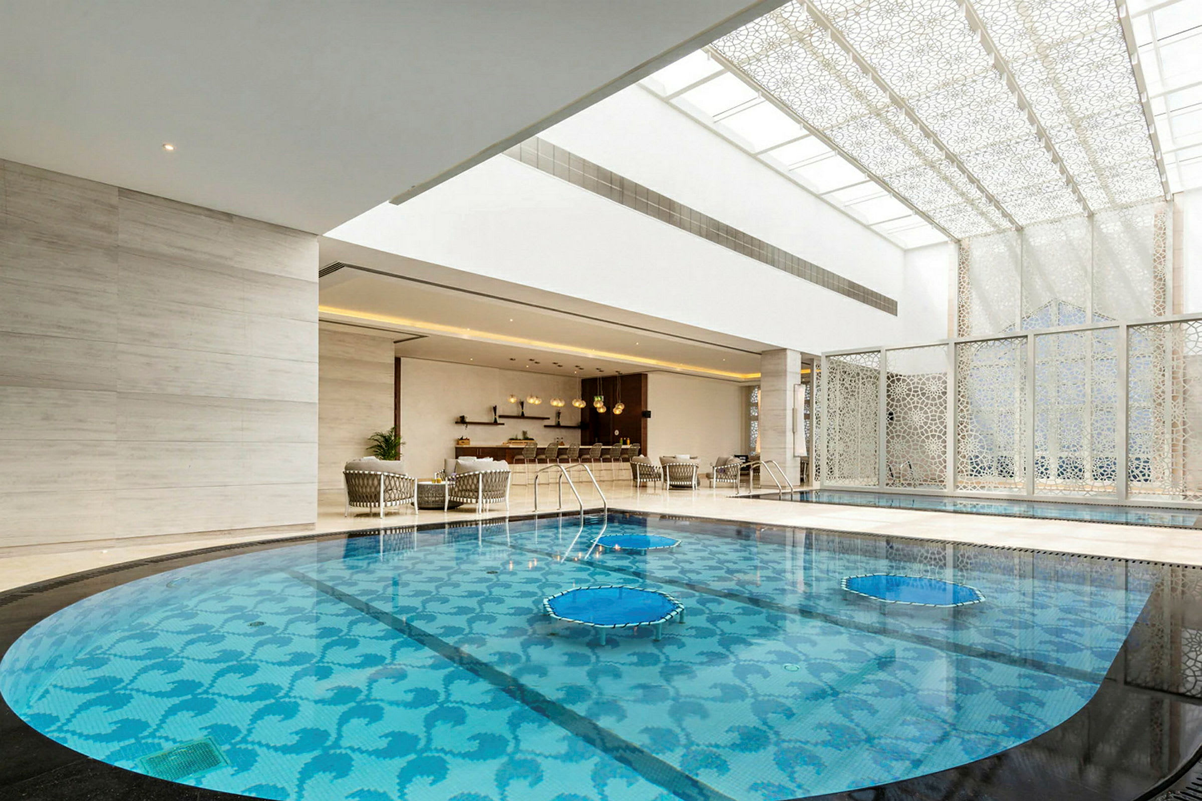 En inomhuspool med ett dekorativt mönster på botten, omgiven av en elegant lounge med sittmöbler och ett tak med stilfullt designade paneler. (AI-genererad text)