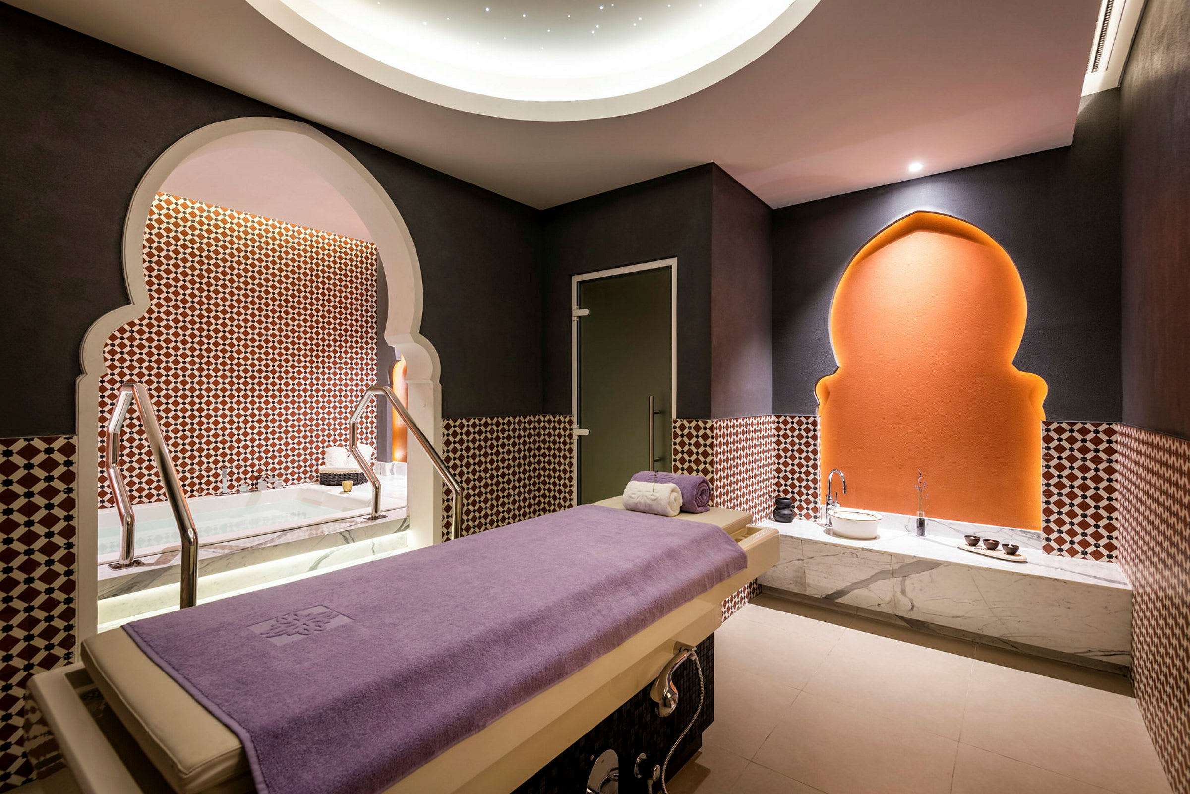 Ett spa-rum med en massagebänk täckt av en lila handduk, dekorativa mönster på väggarna och ett upplyst valv i bakgrunden. (AI-genererad text)