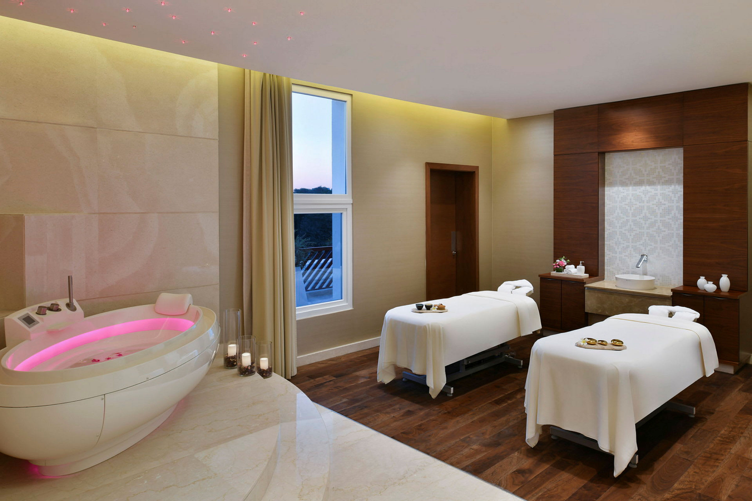 Ett spa-rum med två massagebänkar, ett ovalt badkar med rosa belysning och stora fönster som släpper in ljus. (AI-genererad text)