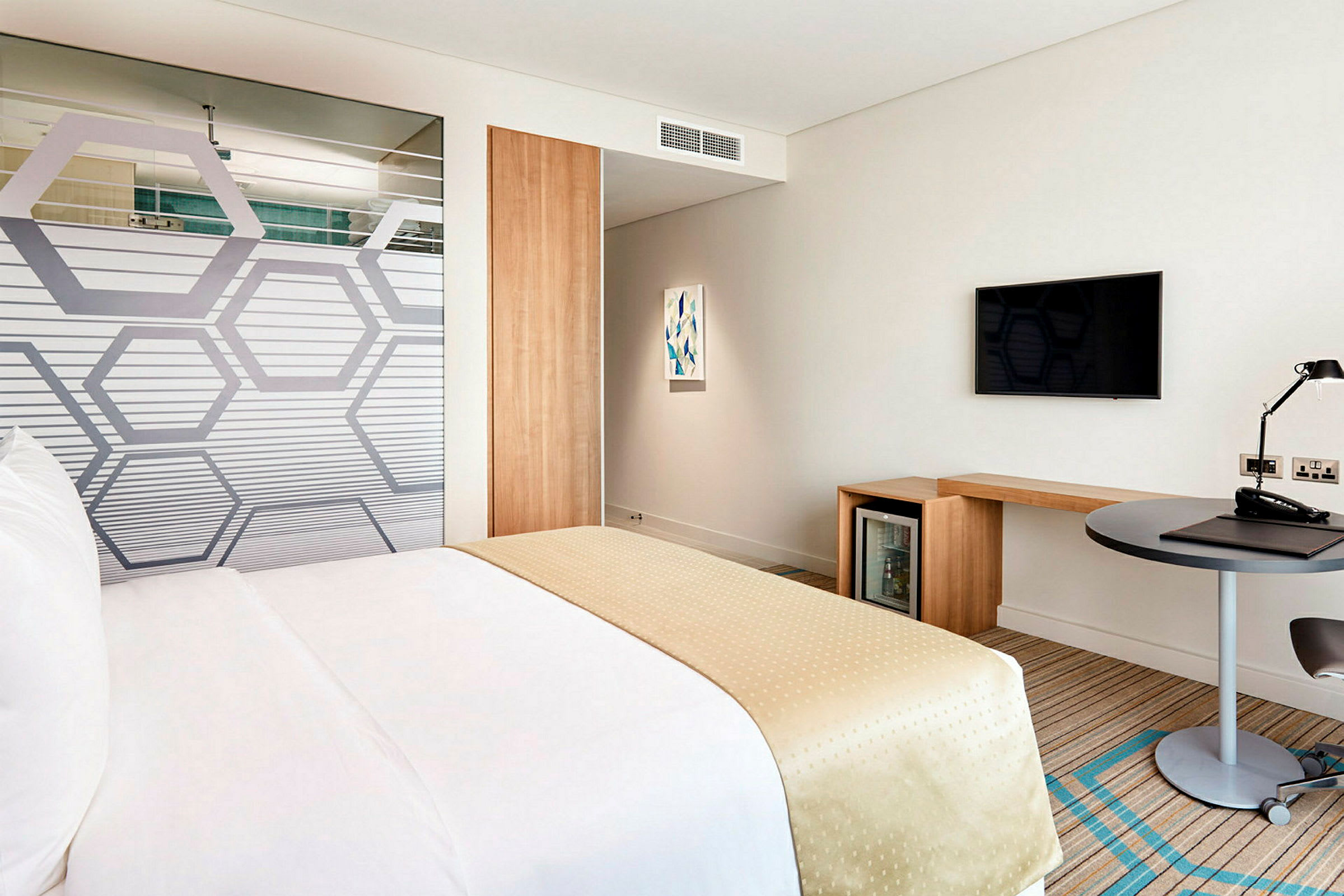 En hotellrum med en säng klädd i vita och beige färger. Rummet innehåller också ett skrivbord, en TV på väggen och ett minikylskåp. (AI-genererad text)