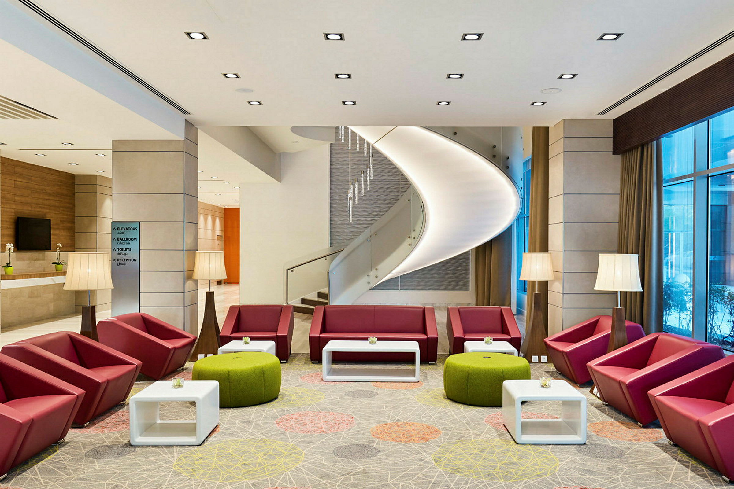 En modern lobby med röda sittmöbler, gröna pallar och vita bord. En spiraltrappa syns i bakgrunden. (AI-genererad text)