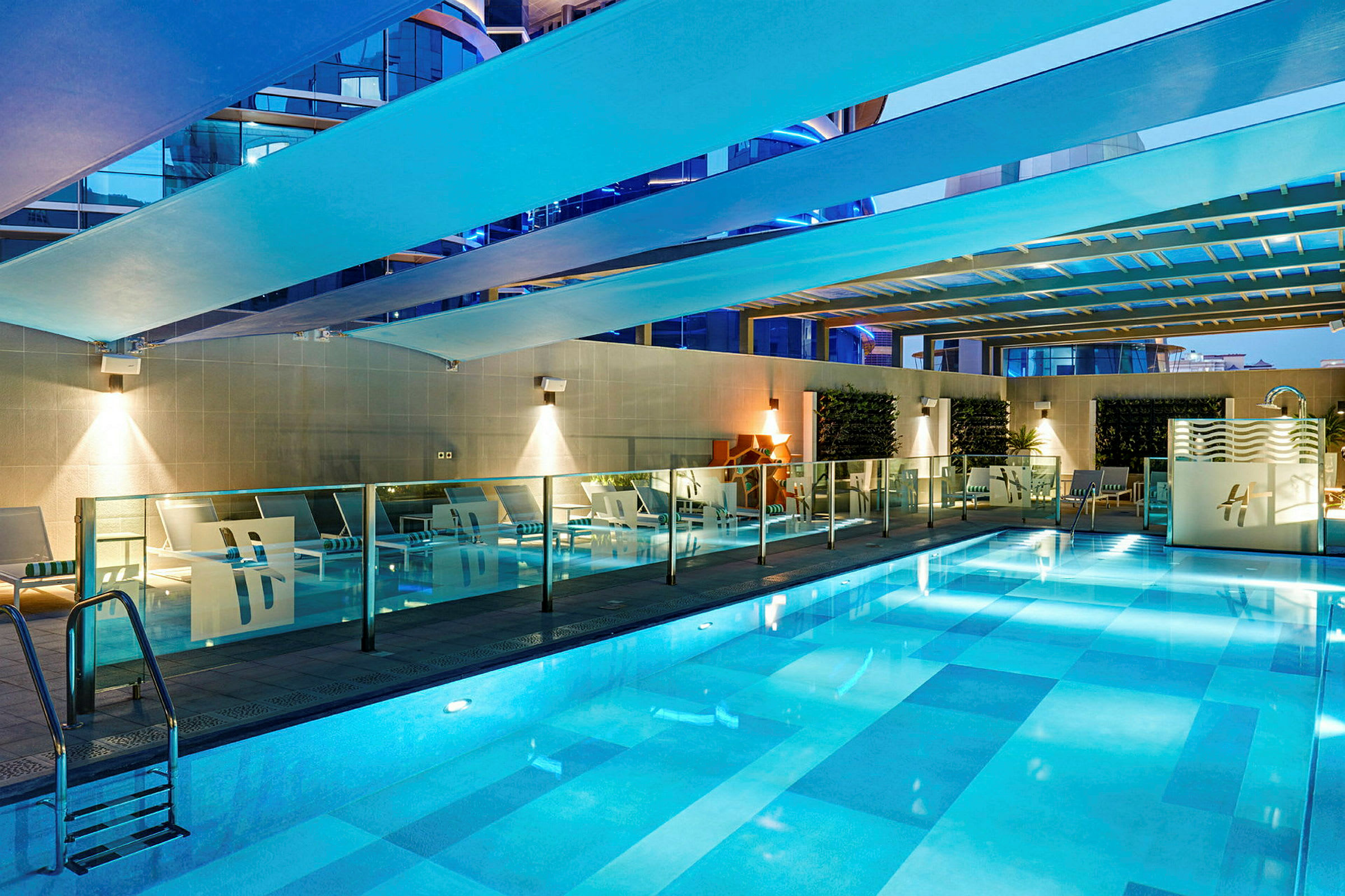 En pool inomhus med belysning. Runt poolen finns vilstolar och glasräcken. (AI-genererad text)