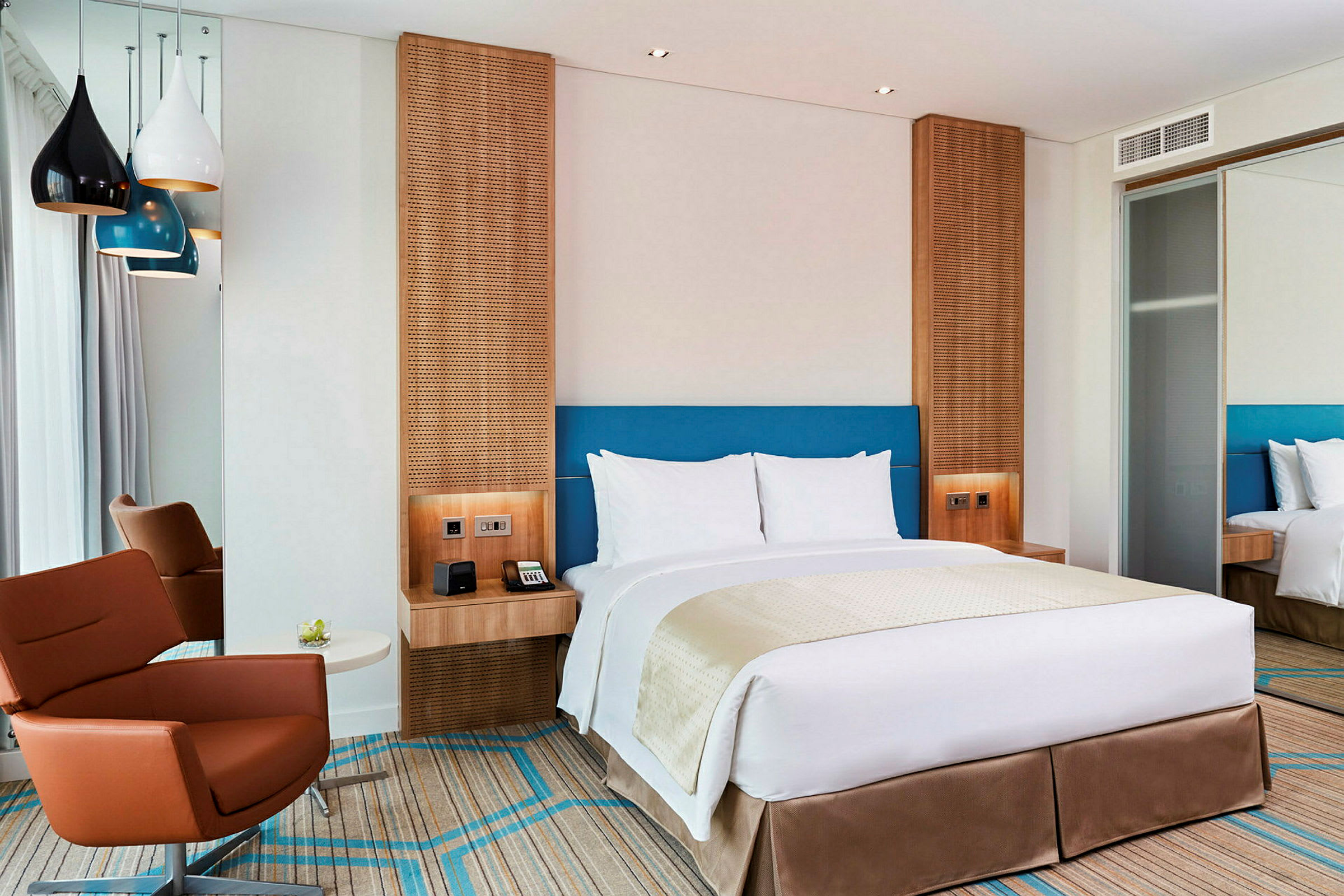 Ett modernt hotellrum med en dubbelsäng, en brun fåtölj och färgglada lampor som hänger från taket. (AI-genererad text)