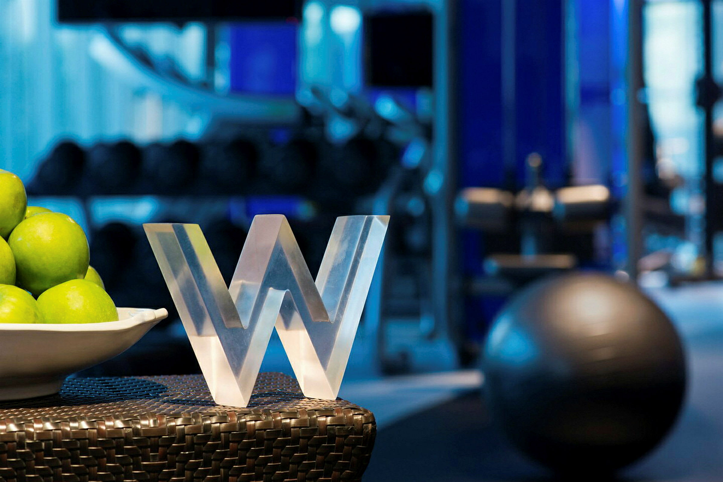 En närbild av en skulptur i form av bokstaven 'W' placerad bredvid en vit skål fylld med gröna äpplen. I bakgrunden syns träningsutrustning på ett gym. (AI-genererad text)