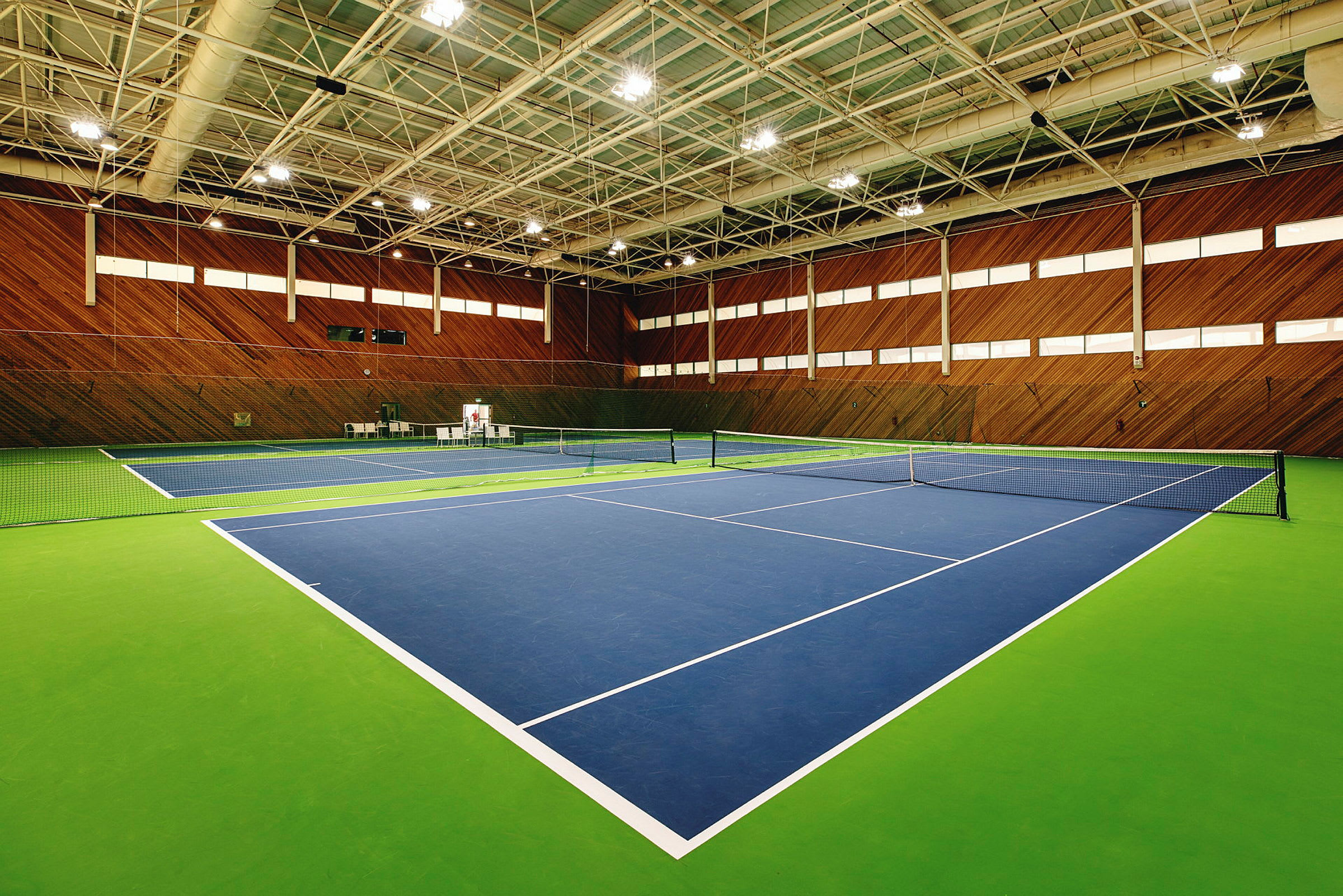 Ett inomhustenniscenter med två tennisbanor, målade i blått och grönt, omgivna av träpaneler på väggarna. (AI-genererad text)