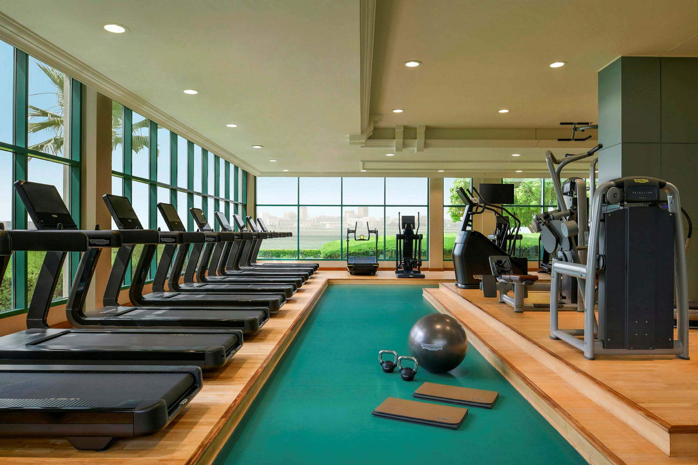 Ett gym med löpband, styrketräningsmaskiner och träningsutrustning. Stora fönster ger utsikt över grönska och stad. (AI-genererad text)