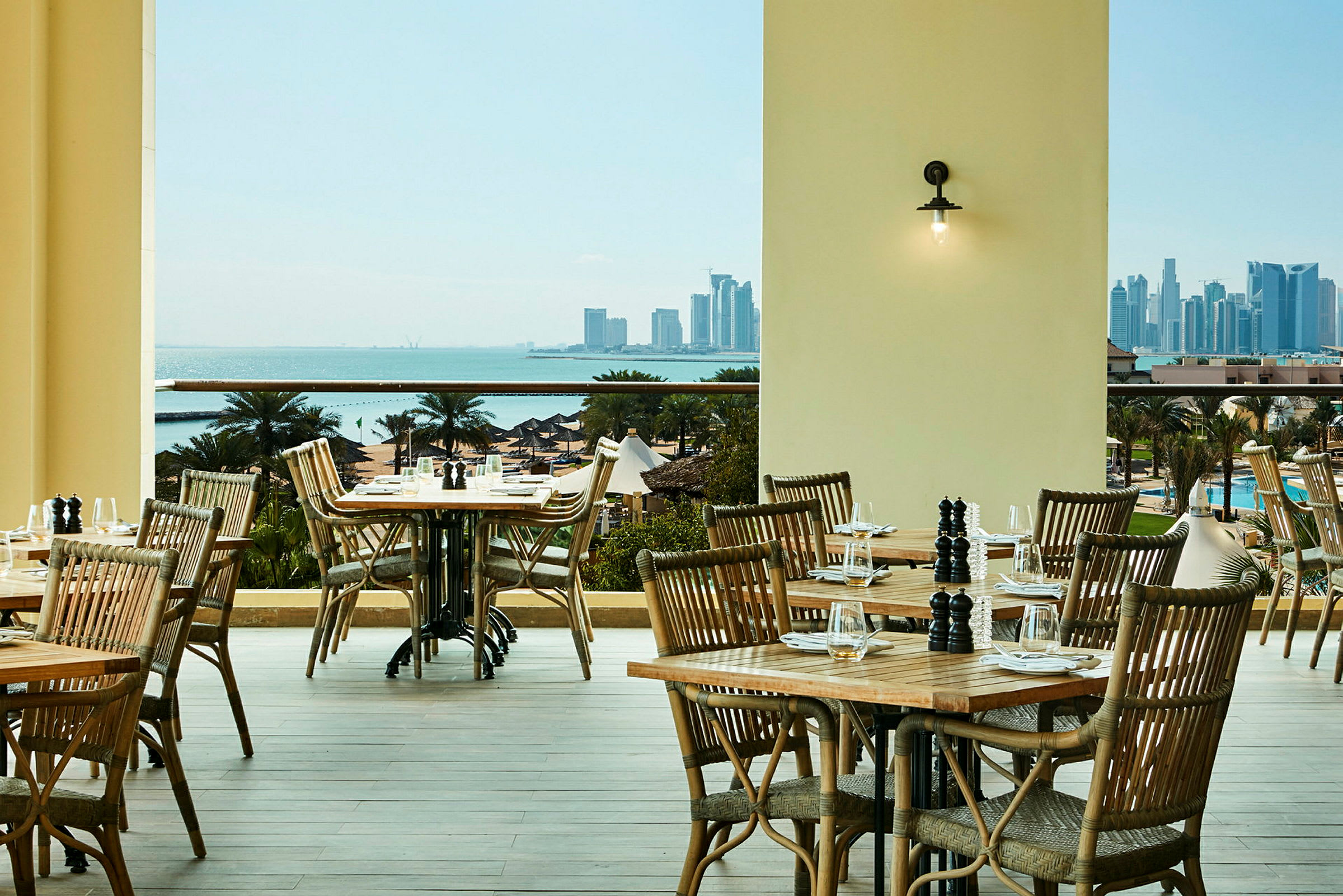 En restaurangterrass med trämöbler som vetter mot en vy över havet och en stadssiluett. (AI-genererad text)