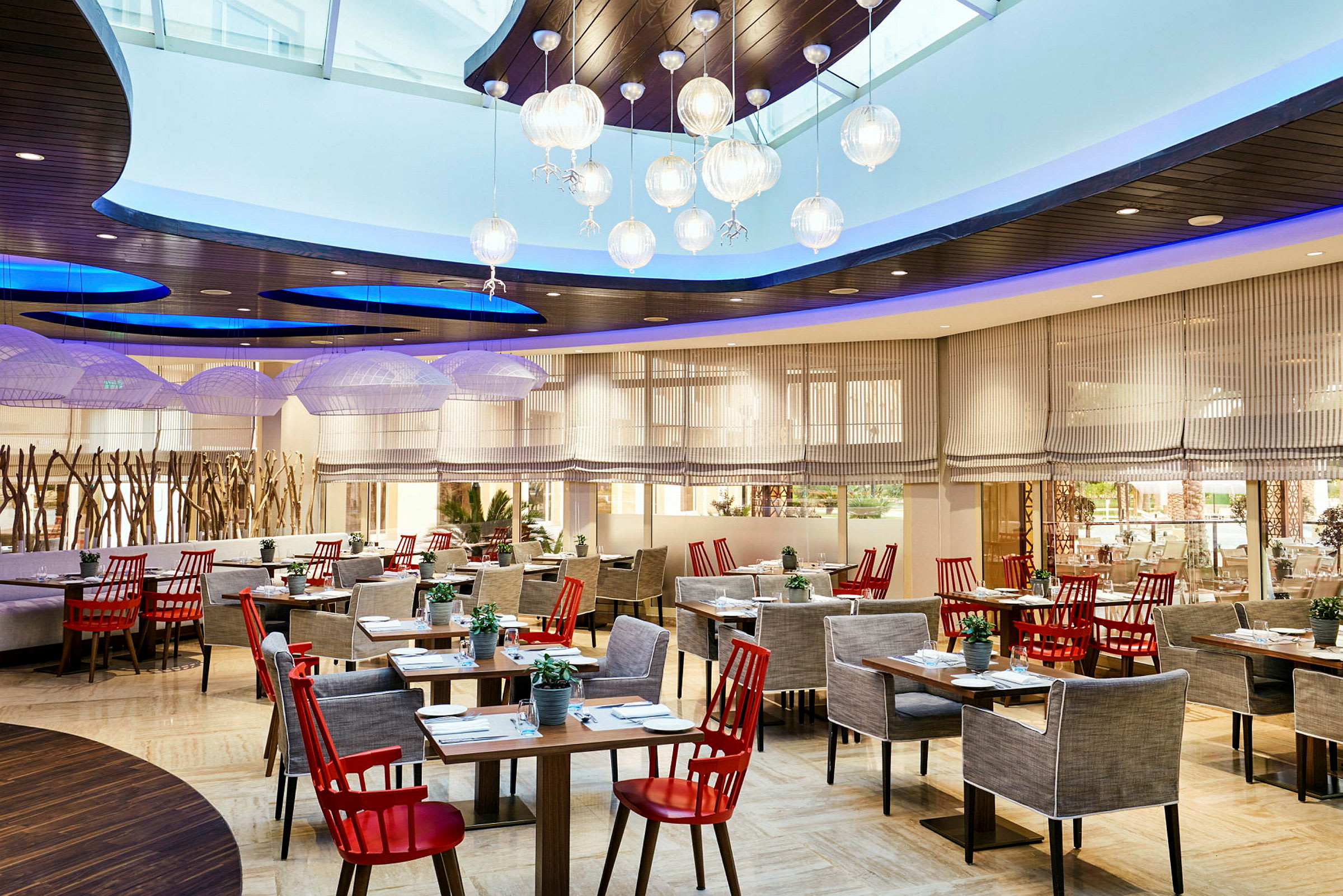 Restaurang med elegant inredning, röda stolar och grå fåtöljer vid flera bord, med moderna lampor i taket och stora fönster. (AI-genererad text)