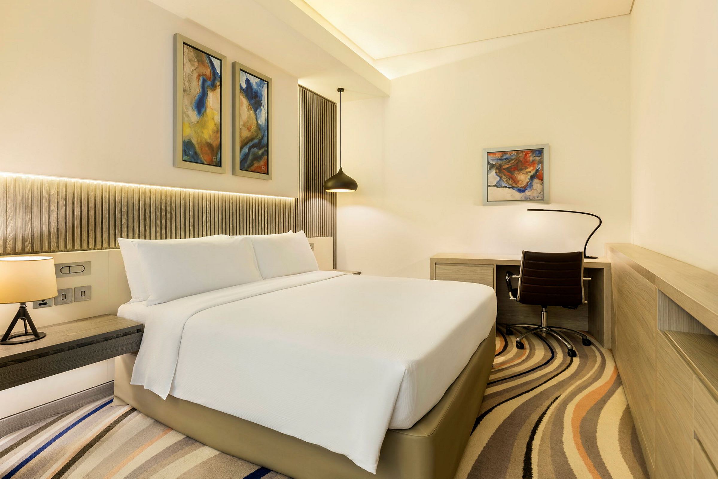Ett modernt hotellrum med en stor säng, skrivbord och stolar samt abstrakt konst på väggarna. (AI-genererad text)