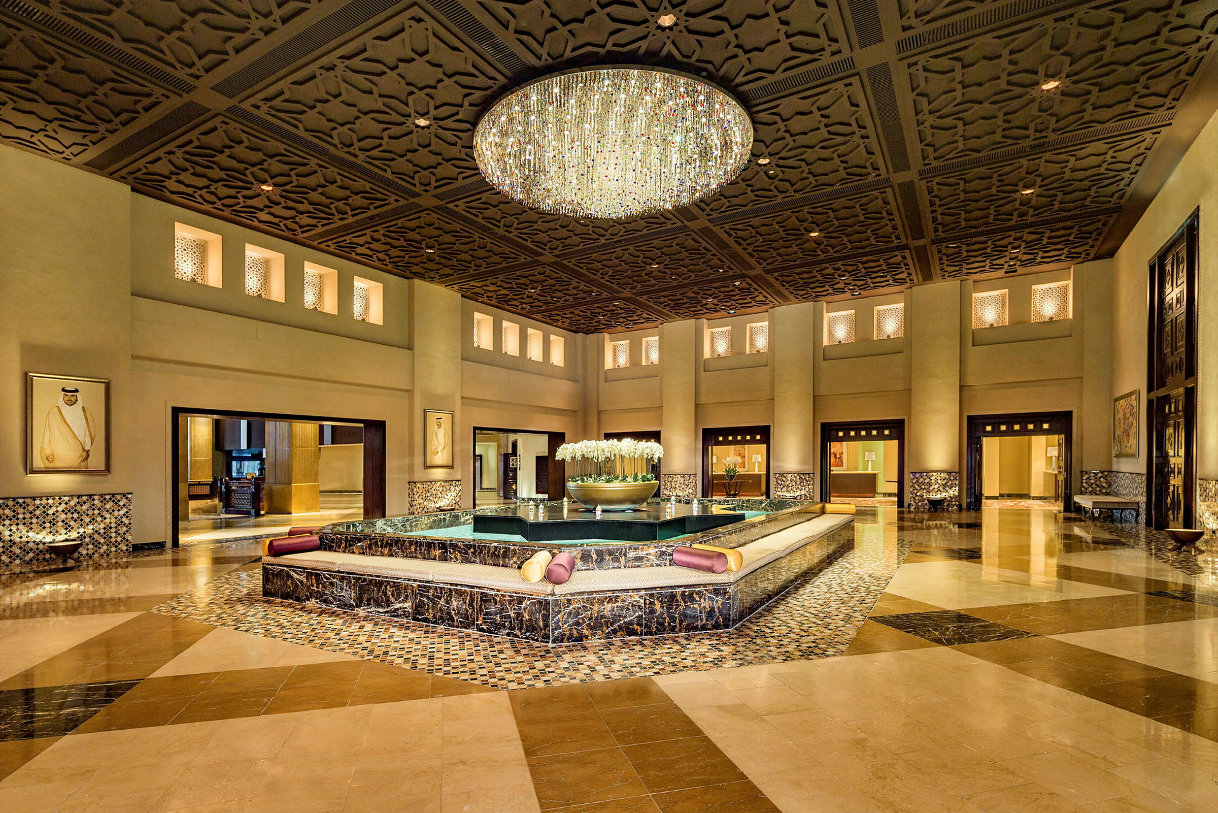 Ett elegant hotellobby med en stor kristallkrona och ett centralt fontänområde omgivet av sittplatser. (AI-genererad text)