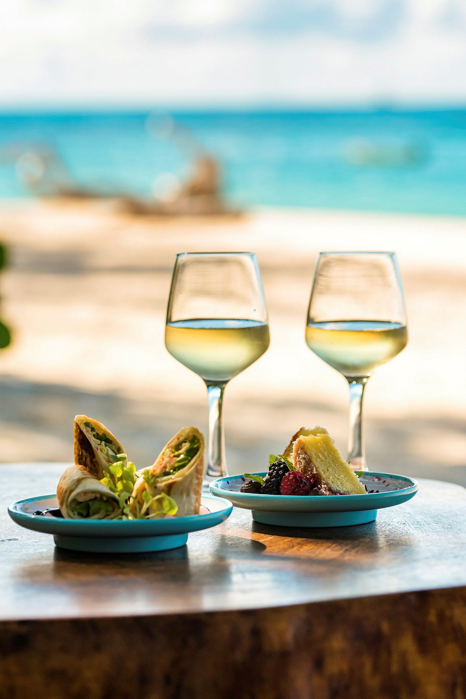 Två vinglas och två tallrikar med mat på ett bord med stranden och havet i bakgrunden. (AI-genererad text)