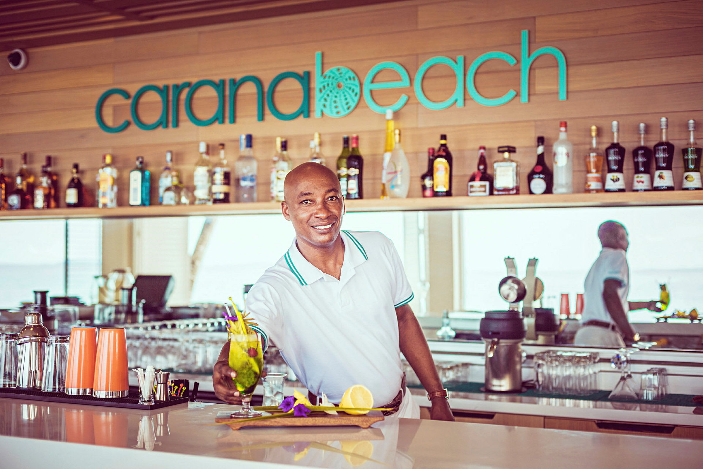 En bartender presenterar en cocktail på en bar med skylt texten 'Carana Beach'. (AI-genererad text)