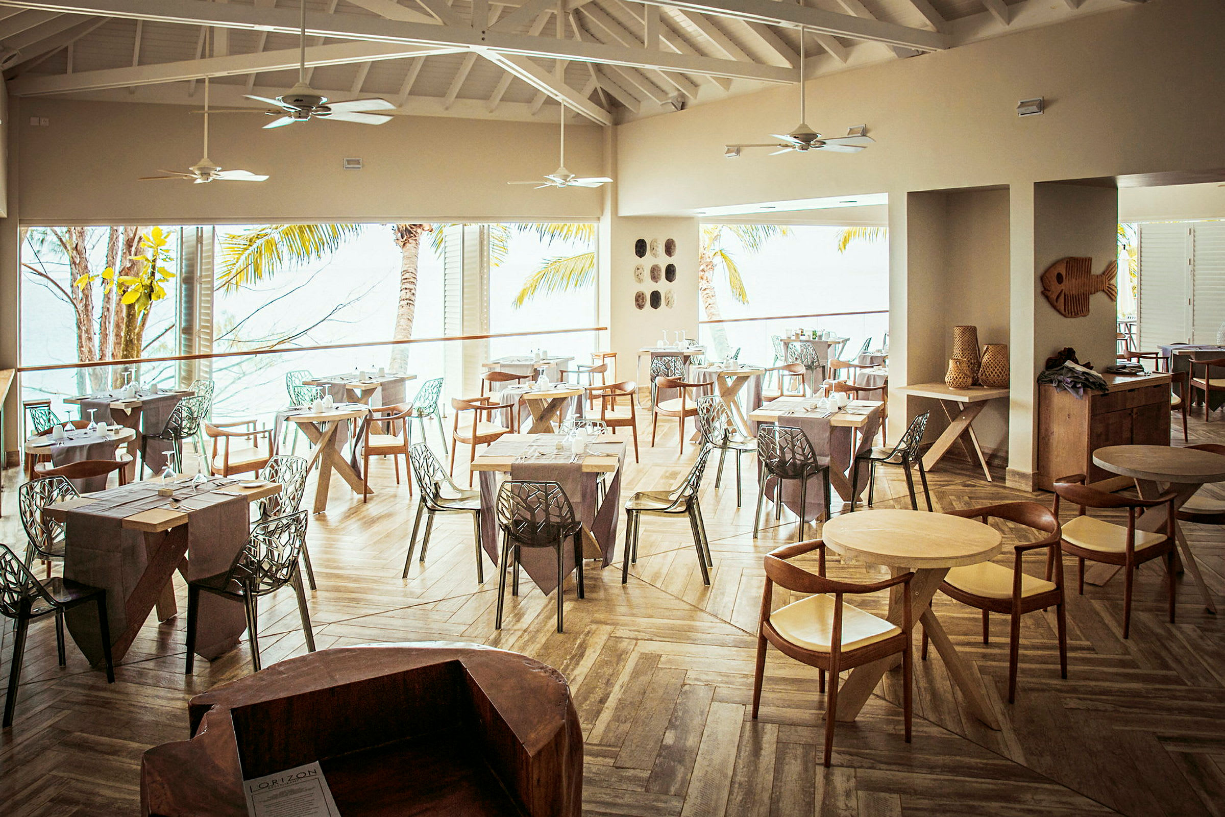 Interiör av en restaurang med träbord och stolar samt stora fönster med utsikt mot palmer och hav. (AI-genererad text)