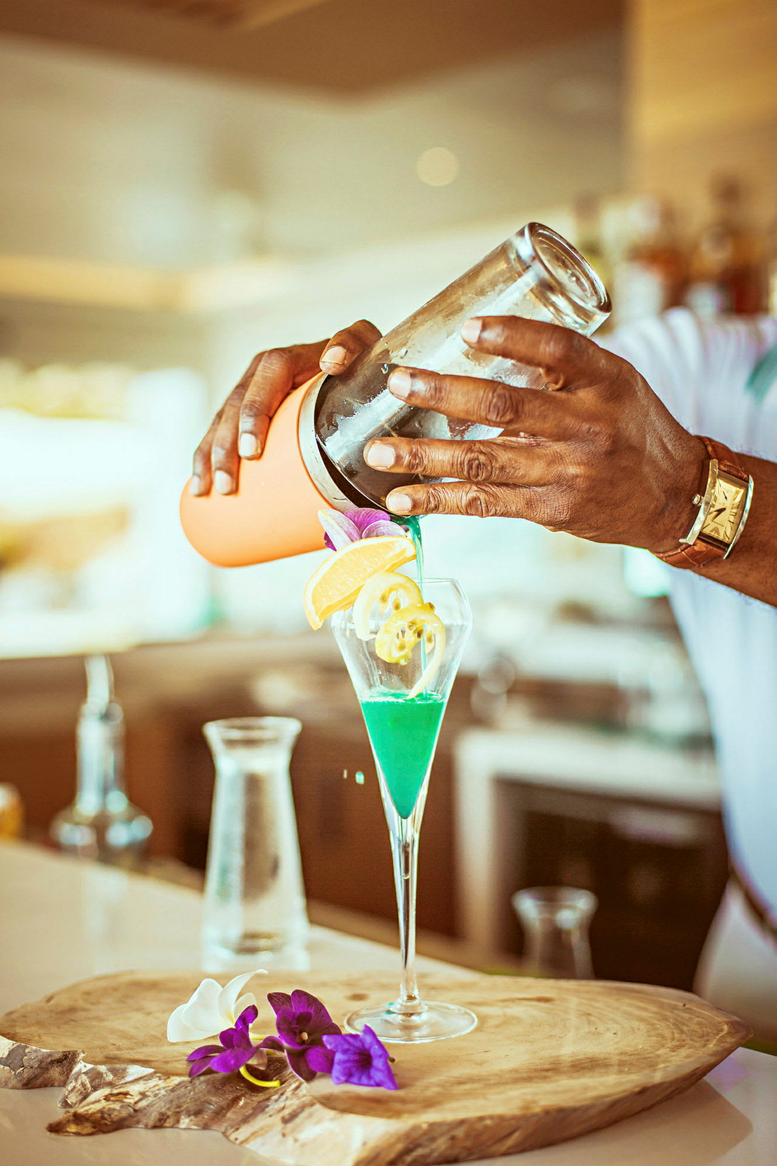 En person häller en grön drink i ett glas med dekorationer som frukt och blommor på en träbricka. (AI-genererad text)