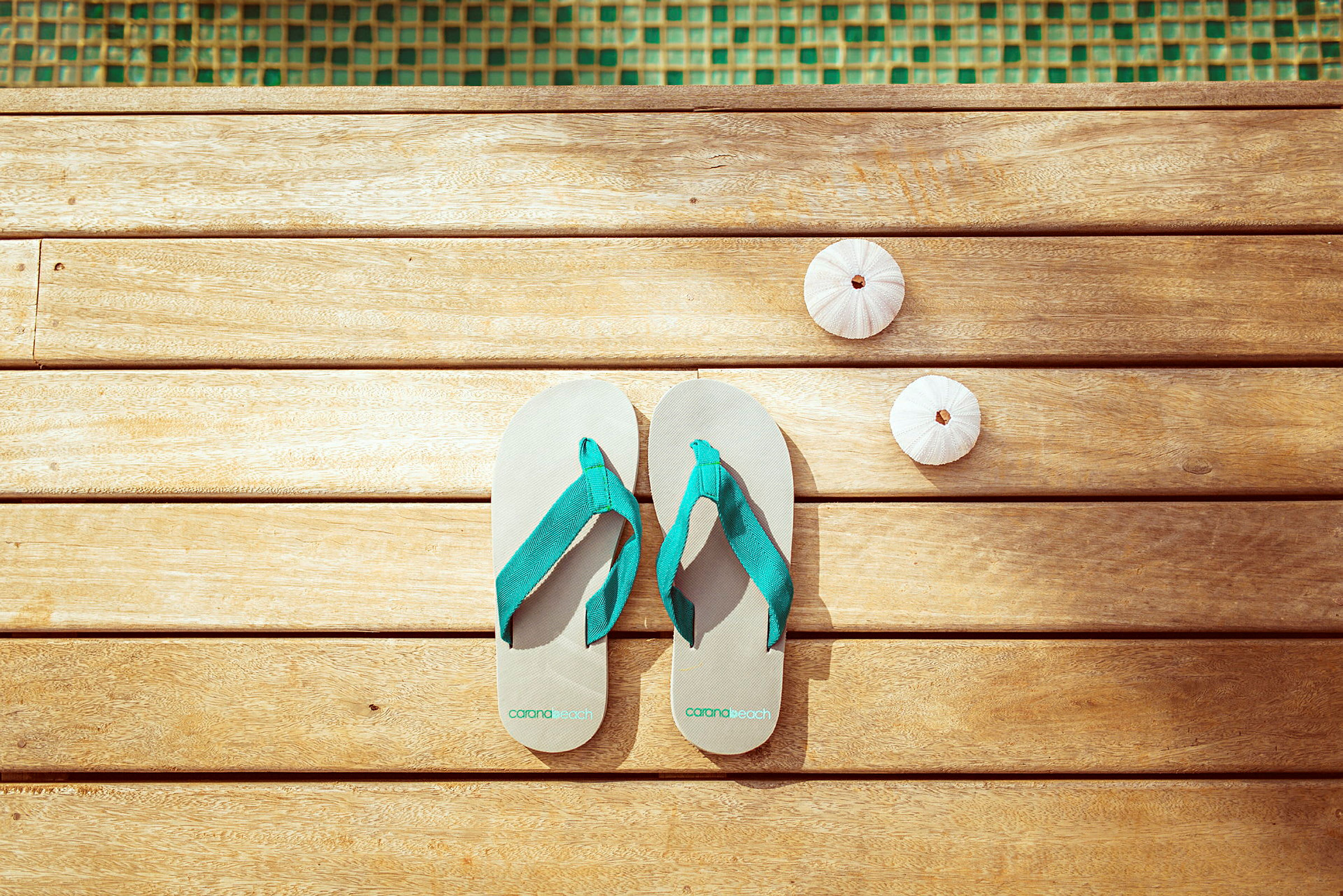 Ett par flip-flops och två små vita snäckskal ligger på en träyta nära en pool. (AI-genererad text)