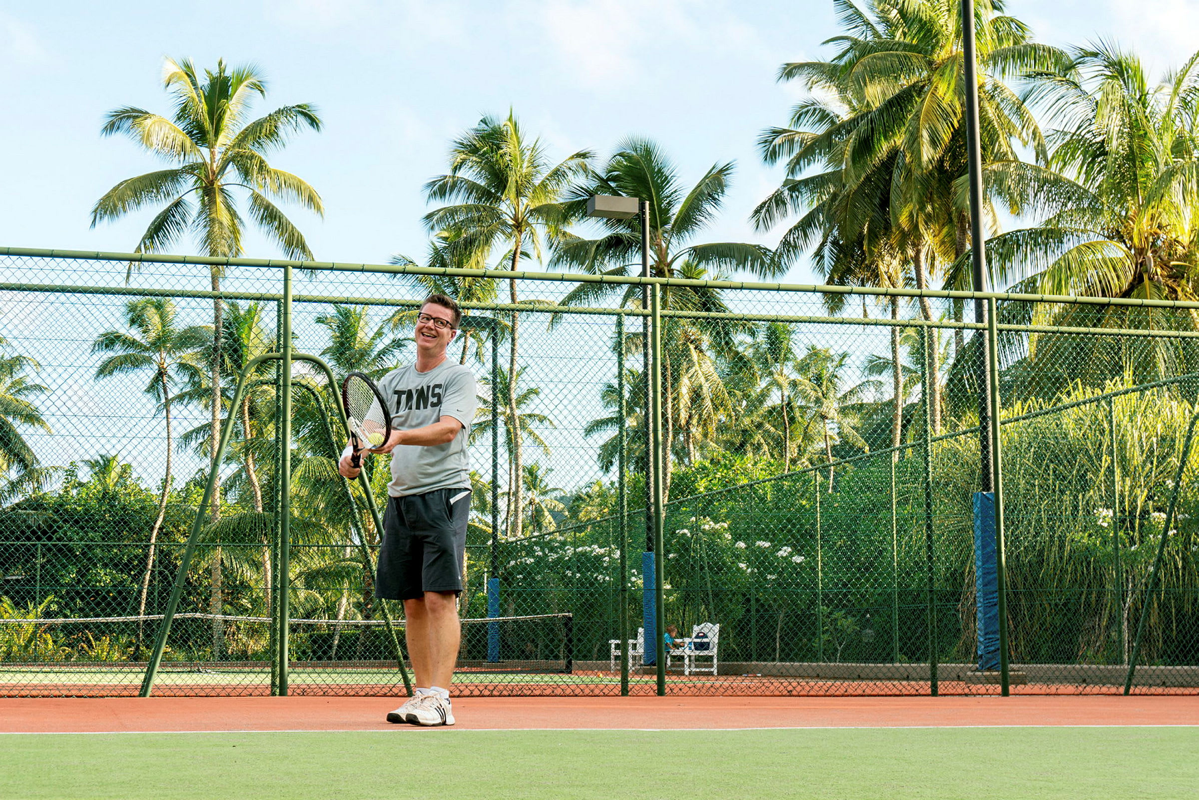 En person spelar tennis på en bana omgiven av grönska och palmer. (AI-genererad text)