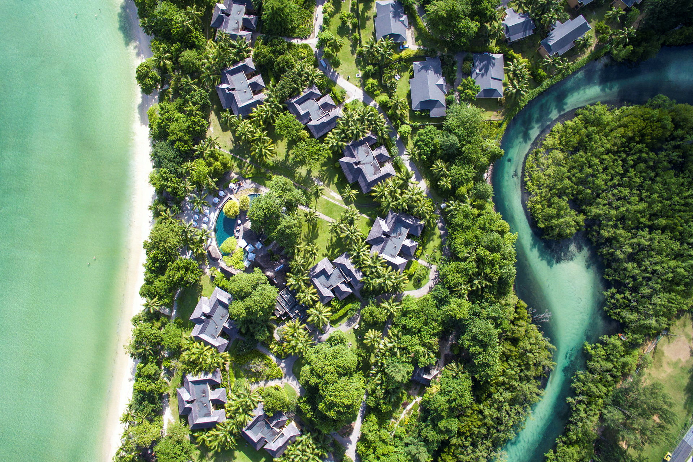 Flygfoto över en tropisk anläggning med byggnader, grönområden, strand och en pool. (AI-genererad text)