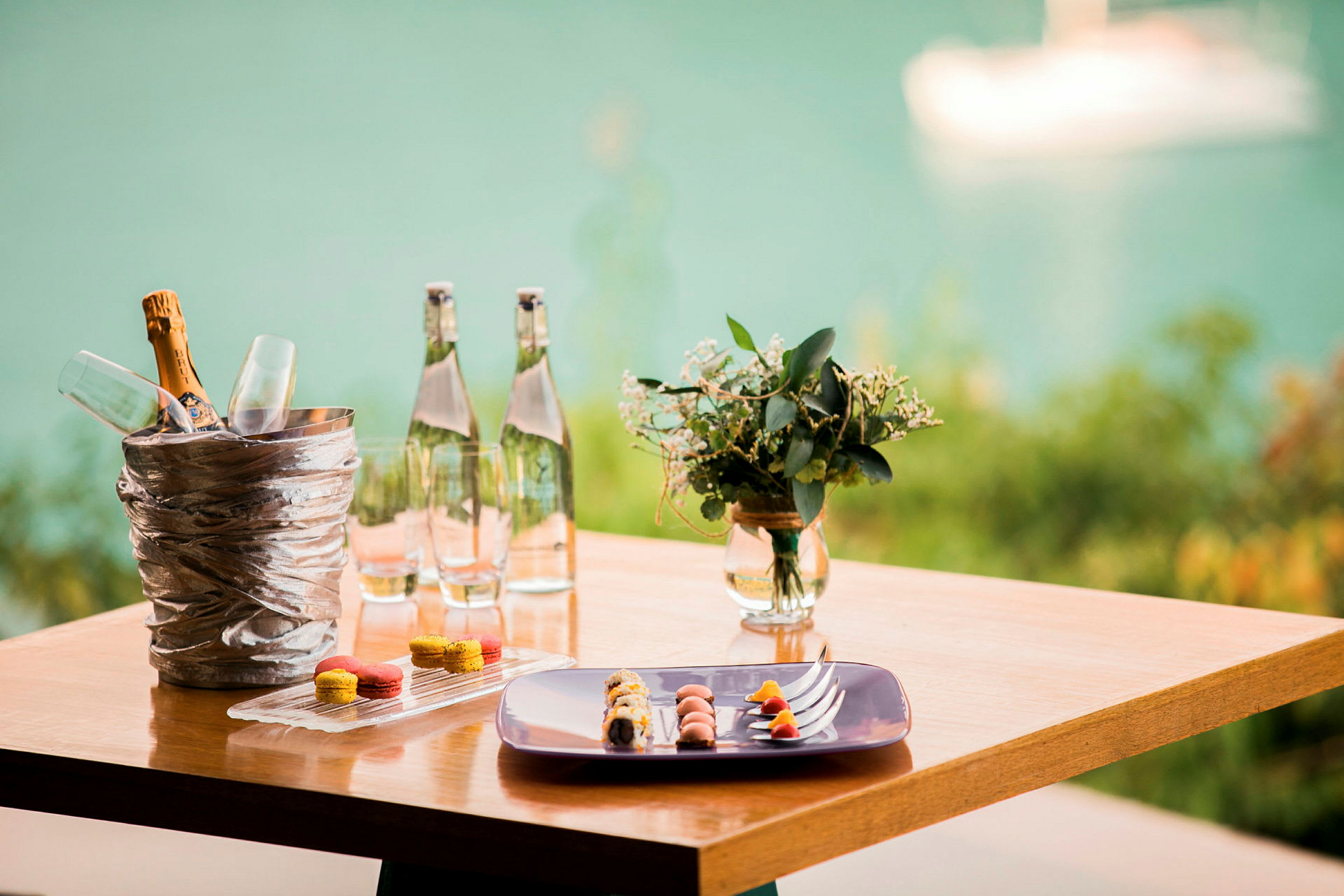 Ett träbord med en hink för champagneflaska och glas, vattenflaskor, en tallrik med mat och små kakor samt en vas med blommor, med en suddig bakgrund av vatten. (AI-genererad text)
