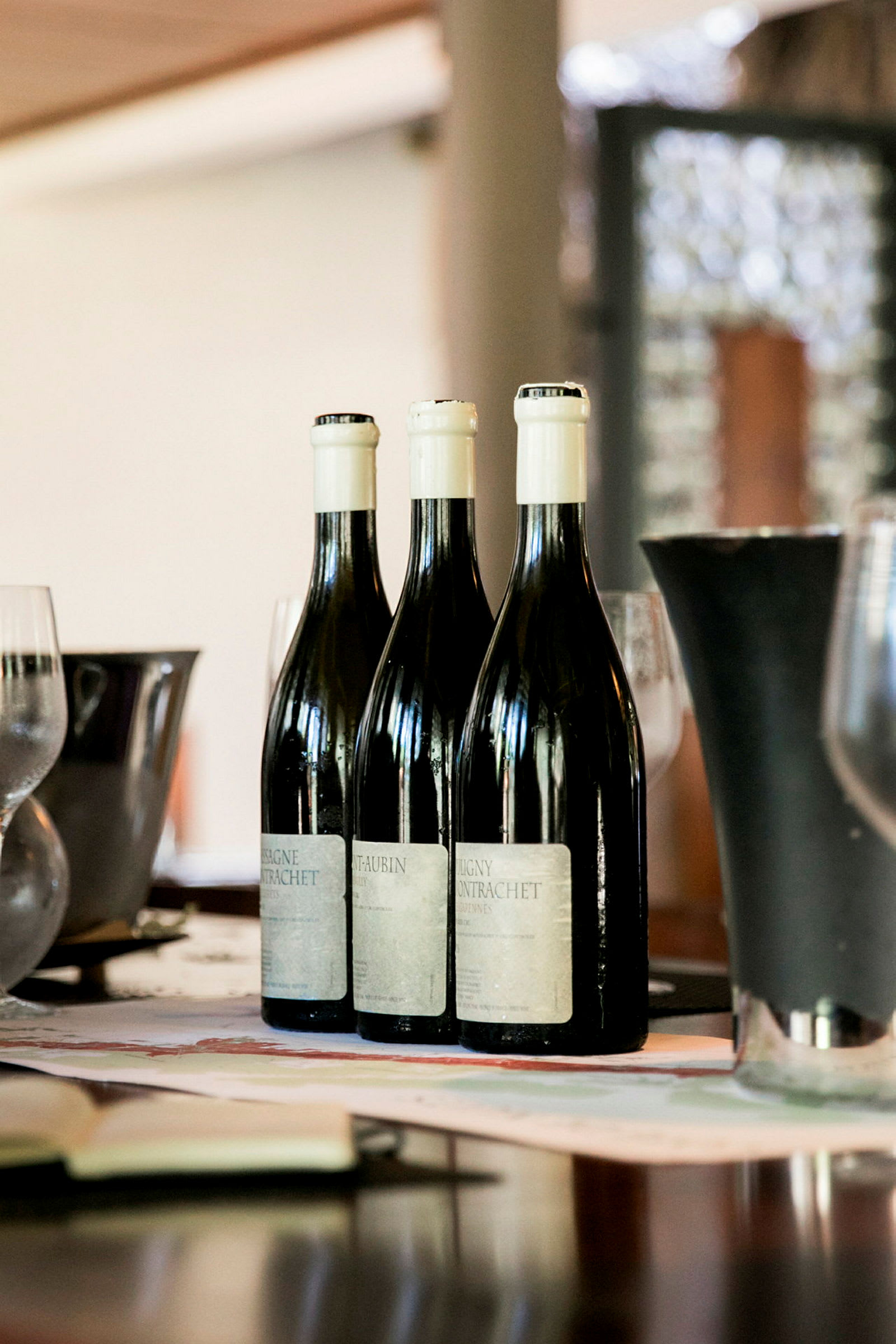 Tre vinflaskor står uppradade på ett bord med glas och en vas i bakgrunden. (AI-genererad text)