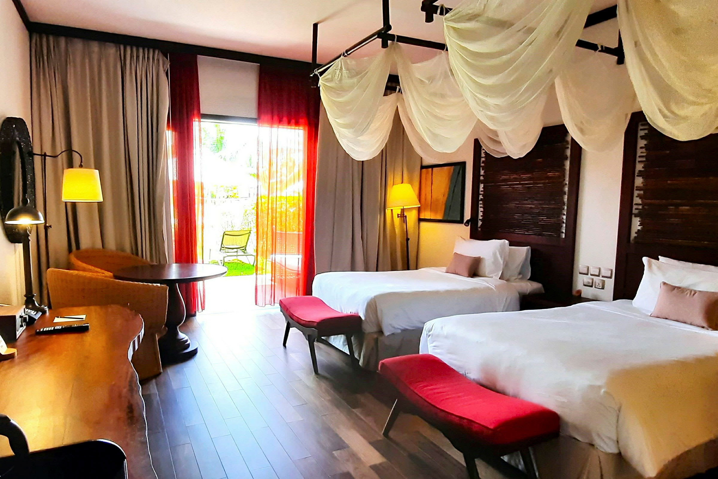 Ett hotellrum med två sängar, vita sängkläder och röda fotpallar. Rummet har dekorativa gardiner, ett skrivbord, en sittgrupp och en balkongdörr som leder ut till en grönskande trädgård. (AI-genererad text)