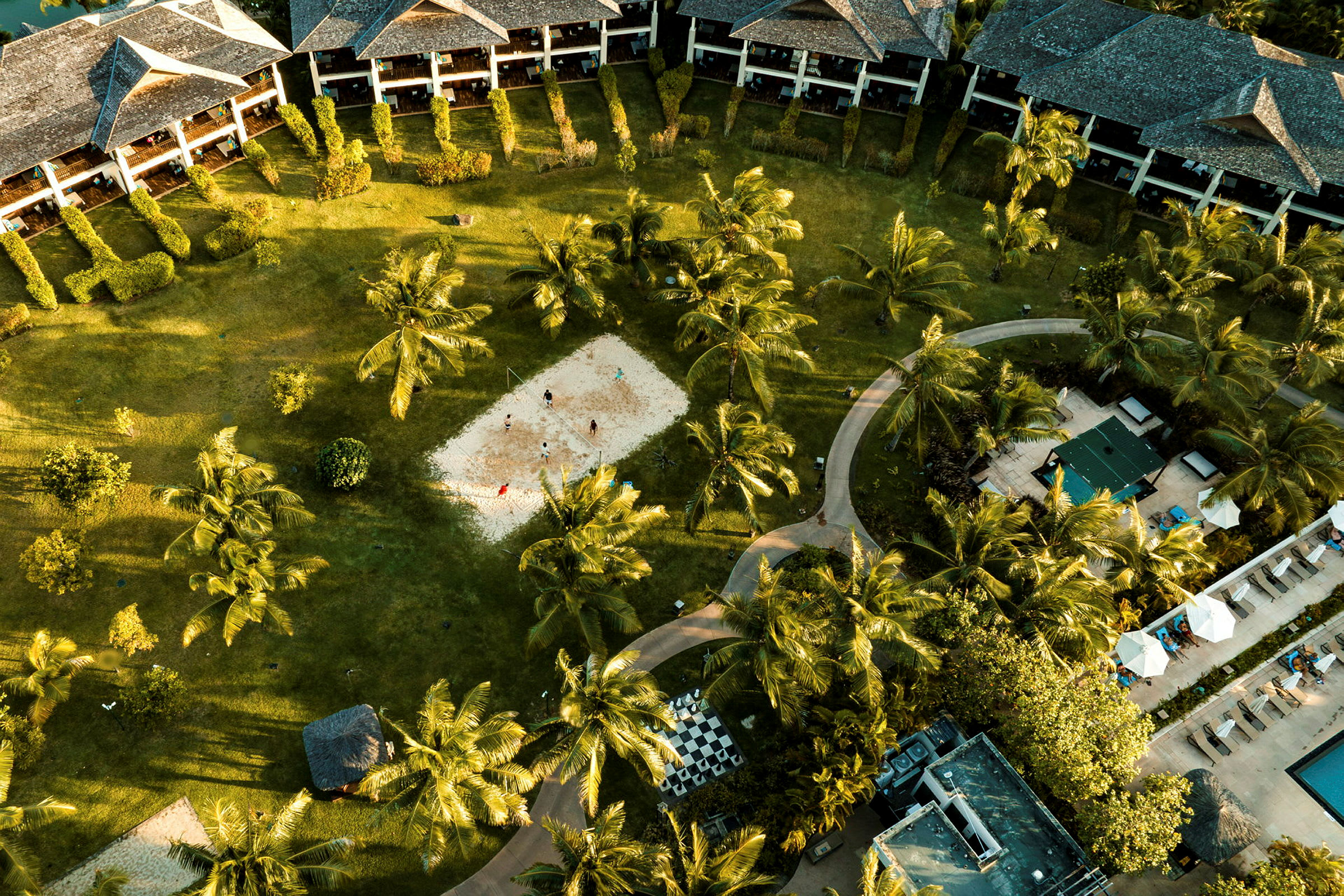 En resortområde med byggnader, grönska, palmer, en sandplan för aktiviteter och en pool- och terrassområde. (AI-genererad text)