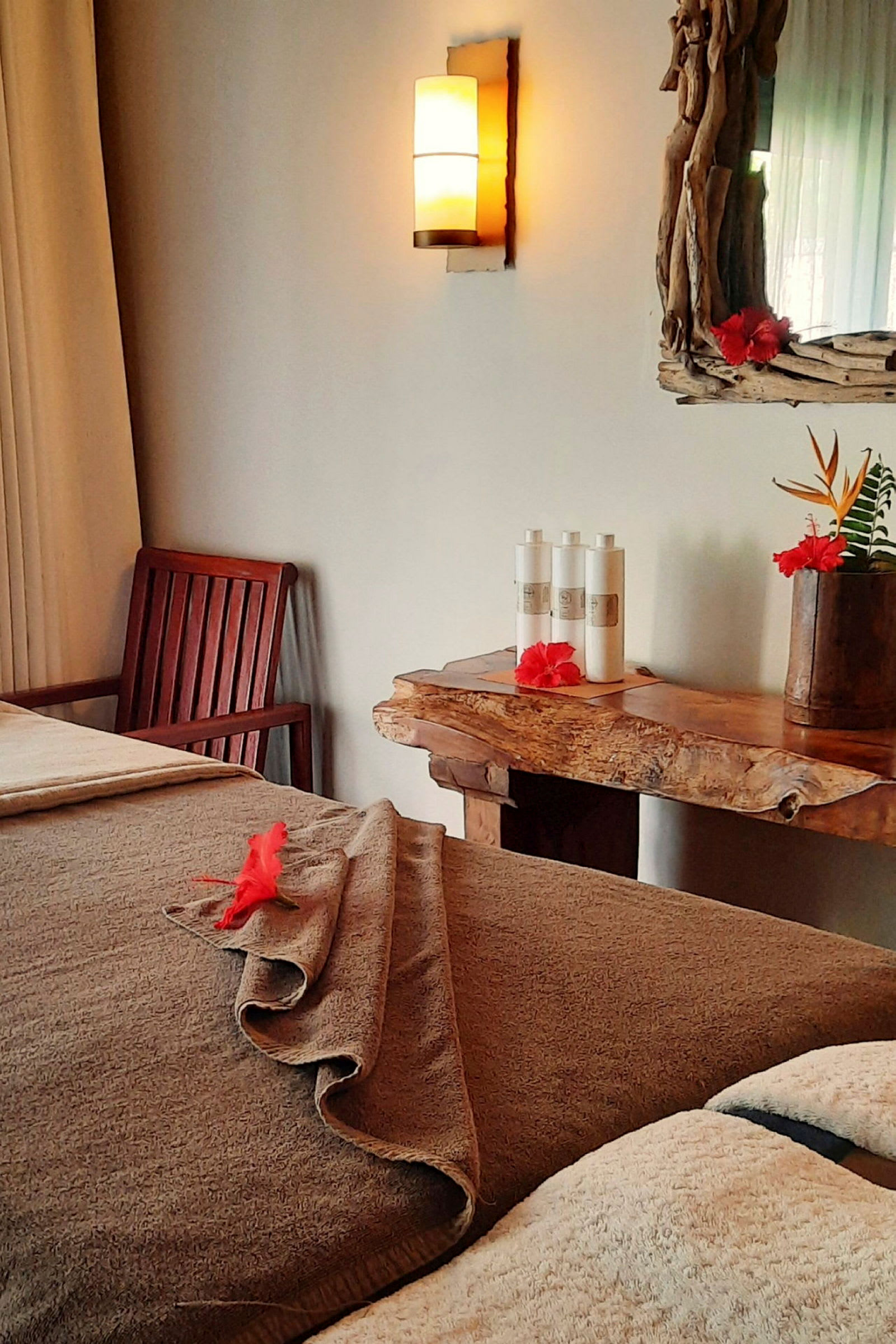 Ett rum med en massagebänk täckt av handdukar och prydd med en röd blomma. En trästol och ett bord med toalettprodukter och en spegel ses i bakgrunden. (AI-genererad text)