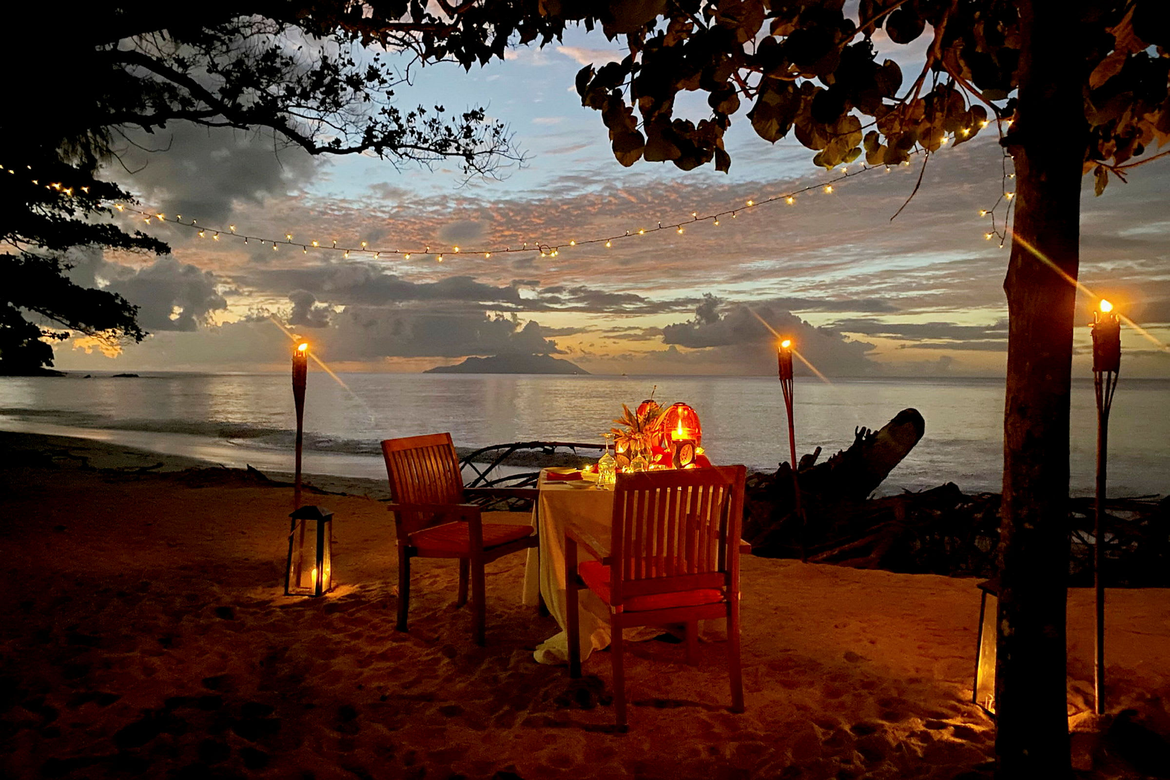 Ett dukat bord med två stolar på en strand, omgivet av lyktor och facklor i solnedgången. (AI-genererad text)