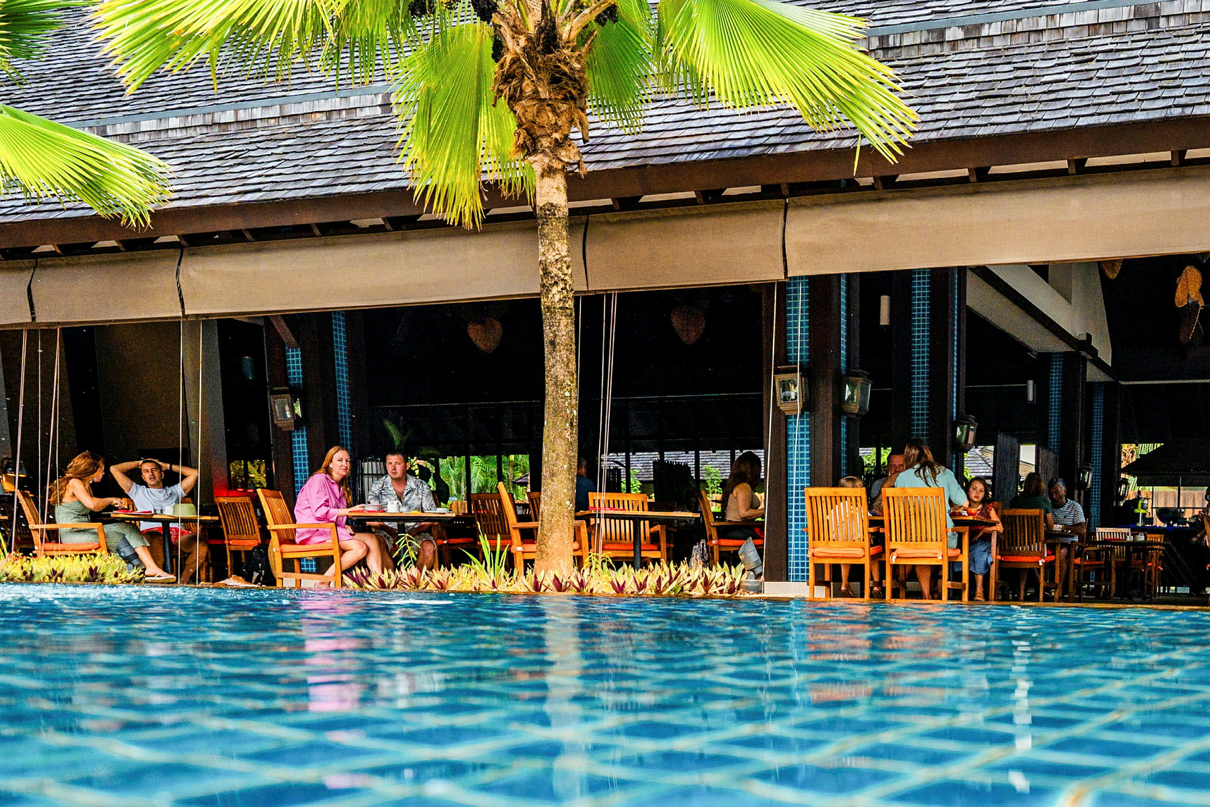 Bild av en restaurang vid en pool, personer sitter vid bord utomhus. (AI-genererad text)