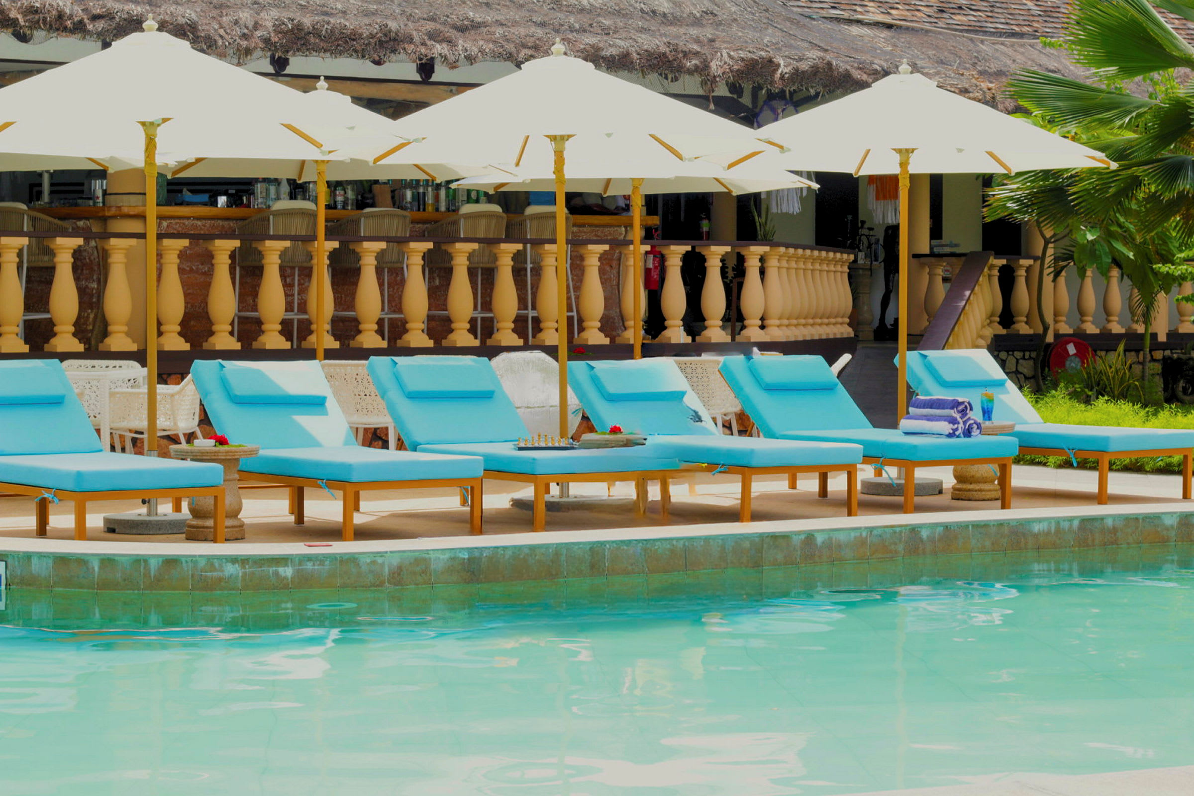 Bild på en pool med flera blå solstolar och parasoller i förgrunden, och en byggnad med träbalustrad i bakgrunden. (AI-genererad text)